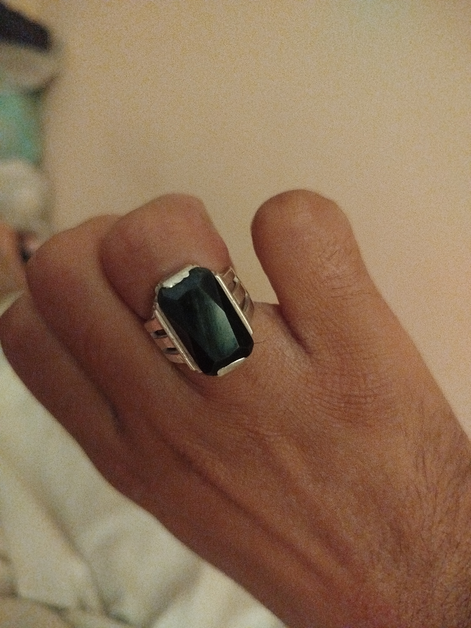 A close-up, overhead shot shows a hand wearing a silver ring with a dark, rectangular gemstone. The hand is positioned in the center of the frame, with the ring on the index finger. The background is a plain, light-colored wall. There is no discernible location in Lassan, Pakistan.