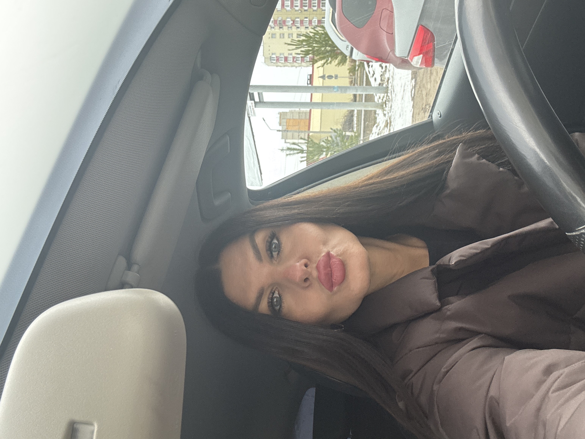A woman is in a car. She is wearing a brown puffer jacket. Outside the car, there is snow and a building that suggests an urban area of Chaikovsky, Russia. The street sign is illegible. Another vehicle is visible in the frame.