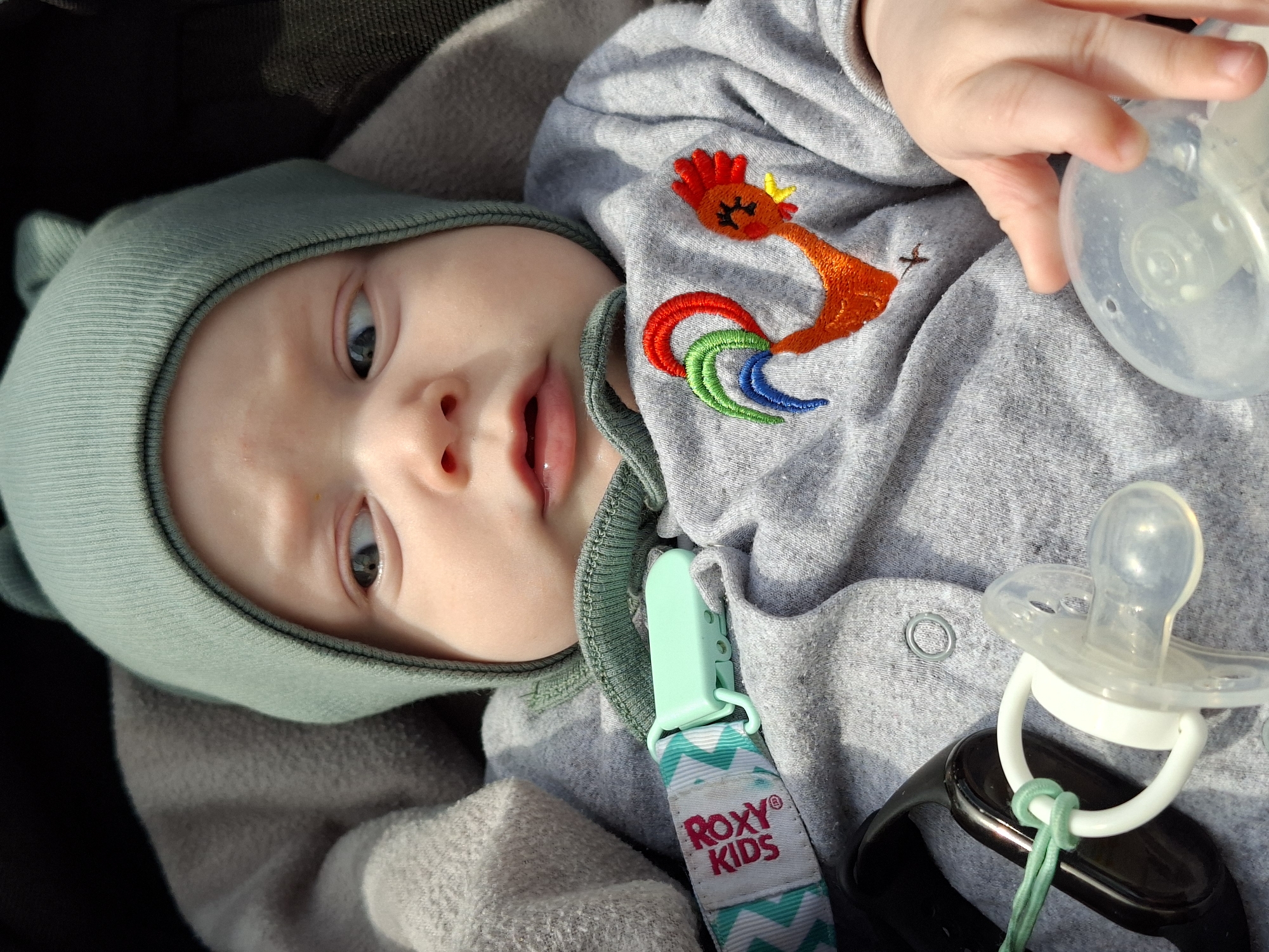 A close-up, high-angle shot depicts a baby wearing a light green beanie and gray onesie with a rooster embroidery. The baby is lying on a grayish blanket. A pacifier and part of a baby bottle are present. A clip with the words "RoXY KIDS" is attached to the onesie. The setting appears to be an indoor or shaded area. The location is not specified in the image or context.
