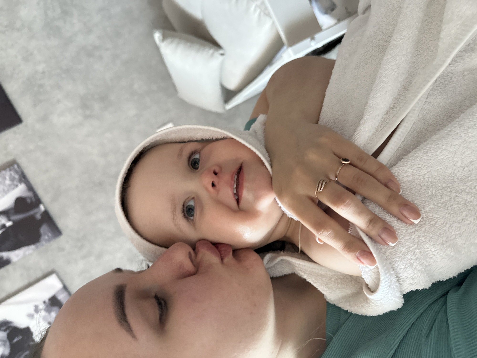 The image captures a close-up, intimate moment between a woman and a young child, likely an infant. The woman's face is turned toward the child, and she appears to be kissing the child on the cheek. The child is wrapped in a white, hooded towel, suggesting they may have recently been bathed. The woman's hand is visible, adorned with multiple rings. The background includes a glimpse of a white couch with pillows and some black and white photos. The location in the city of Chaikovsky, Russia, cannot be confirmed from the provided image.