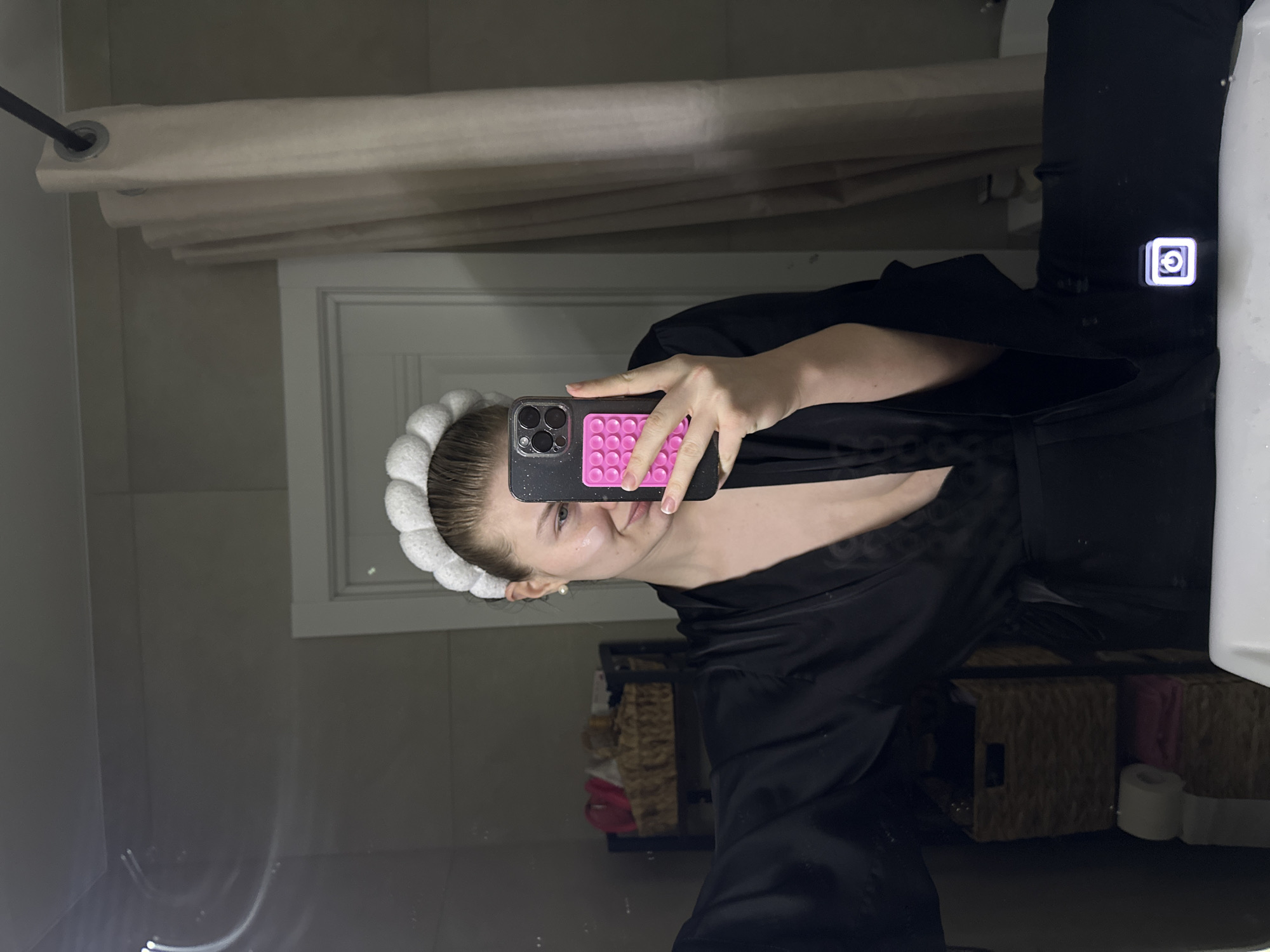 An adult female is depicted taking a mirror selfie in a bathroom in Kazan, Russia. She wears a white, textured headband and a dark, possibly black, robe. A pearl earring is visible in her left ear, and a small amount of white cosmetic product is present on her left cheek. She holds a smartphone with a black case and a pink grid-patterned accessory on the back, partially obscuring the lower half of her face. The reflection in the mirror reveals a light-colored shower curtain on a dark rod to the right, and a white door frame behind it. To the left, a dark-framed shelf unit holds two wicker baskets, one containing a pink item, and two rolls of toilet paper (one white, one grey) stacked below. A white sink basin is visible in the lower foreground, reflecting an illuminated square power symbol, suggesting a smart mirror feature. The walls are light-colored.