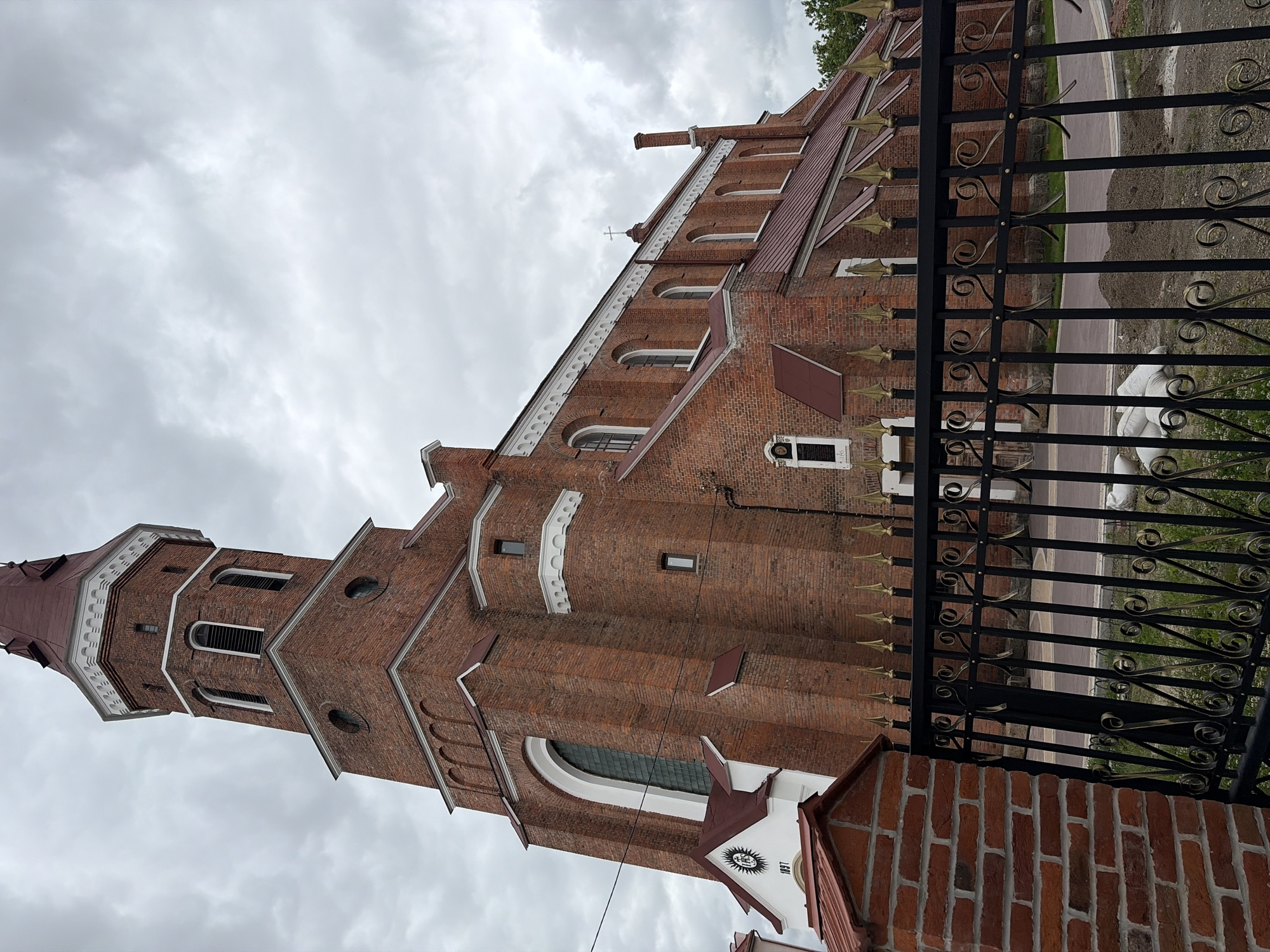This is a low-angle, full shot of the Holy Resurrection Cathedral in Kolomyia, Ukraine, captured on an overcast day. The red brick cathedral is the primary subject, with its tall bell tower dominating the left side of the frame. The tower features arched windows and a white decorative cornice beneath its red, multi-tiered roof. The main body of the church extends to the right, also constructed of red brick with a red roof. Numerous arched windows are visible along its side, with a smaller cross atop the roof. A white facade with a circular emblem and the year "1897" is attached to the lower portion of the cathedral near the bell tower. In the foreground, a black wrought-iron fence with gold accents partially obstructs the view of the cathedral and the paved pathway behind it. A brick pillar stands to the left of the fence. Piles of soil and several white sacks are visible on the ground behind the fence, suggesting ongoing construction or landscaping work. The sky is heavily clouded, casting a diffused light on the scene.