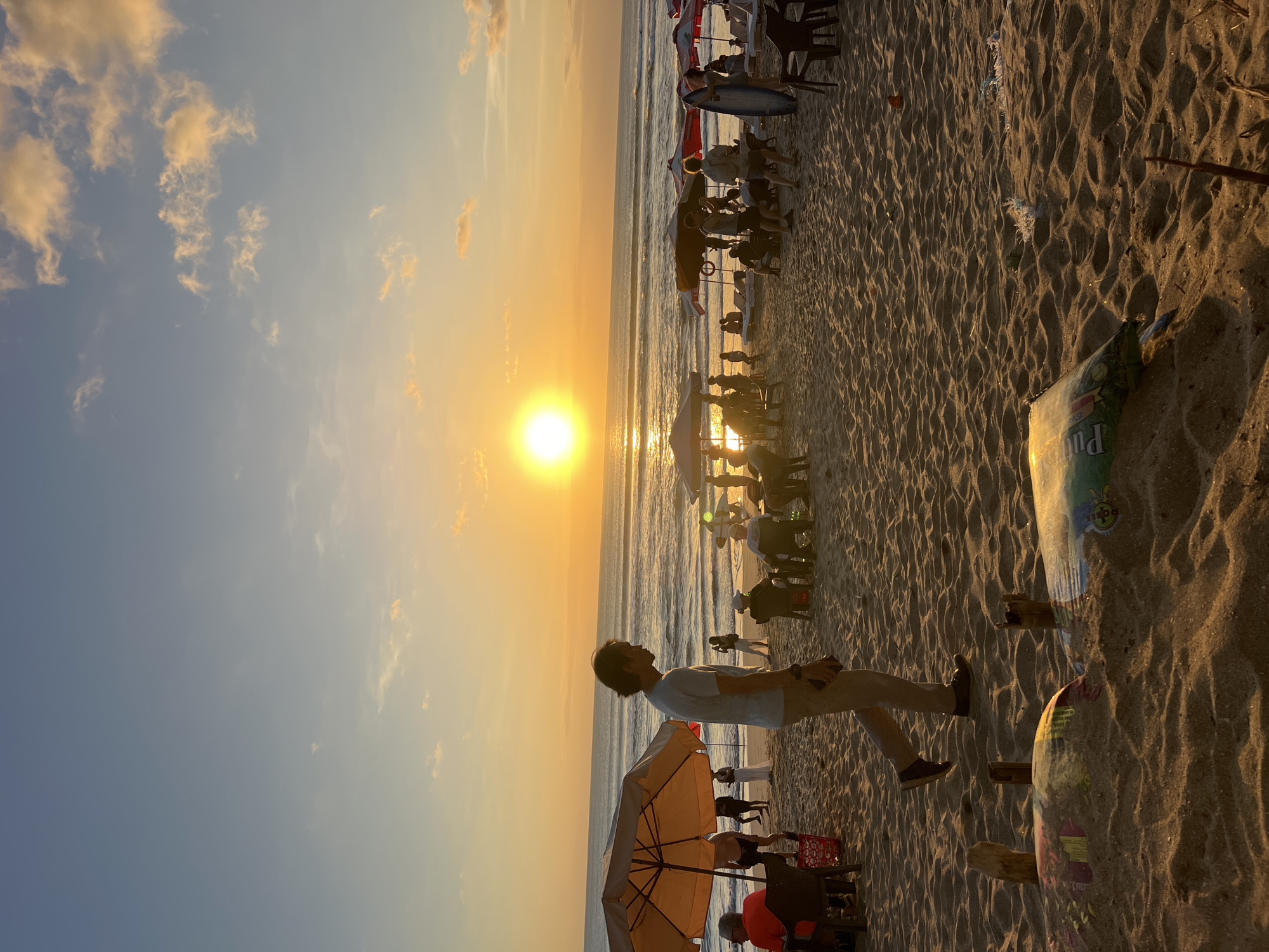 This is a photograph taken at sunset on Kuta Beach, Bali, Indonesia. The sun is a bright orb low on the horizon, casting a golden glow over the ocean and the sandy beach. Several beach umbrellas are set up, some with people seated beneath them. In the foreground, a man is walking along the beach, his profile visible against the setting sun. There are other individuals scattered across the beach, some standing, some sitting, and some walking. The sand is textured with ripples, and some debris is visible on the sand. The waves are gentle, rolling onto the shore under a sky transitioning from blue to orange and yellow hues.