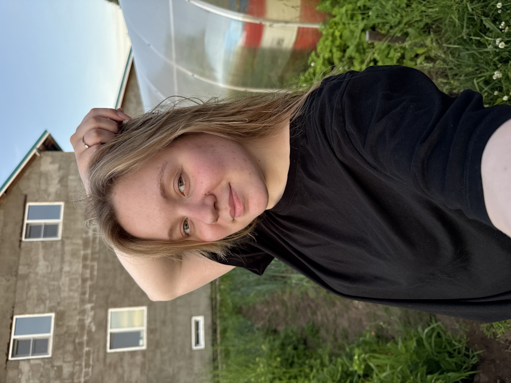 A young Caucasian woman with blonde hair and light green eyes is captured in a close-up, outdoor, selfie-style photograph. She is positioned in the foreground, facing the camera with a slight smile. Her right arm is raised, with her hand resting on the back of her head, and she is wearing a simple black t-shirt. A ring with a small stone is visible on her right ring finger. The background suggests a rural or semi-rural setting. To the left of the woman, a building with multiple windows and rough concrete or stucco siding is visible. The building appears to be under construction or in a state of disrepair. To the right of the woman, a large, transparent greenhouse structure is present, containing red and white cylindrical objects, possibly barrels. The ground in the middle and left foreground is covered with green foliage, with a path of dark soil visible between the plants. The sky above is a clear, pale blue, indicative of daylight, likely late afternoon or early evening given the lighting. The location is identified as Garevaya, Russia, a small settlement known for its agricultural activities, which aligns with the presence of the greenhouse and cultivated land.