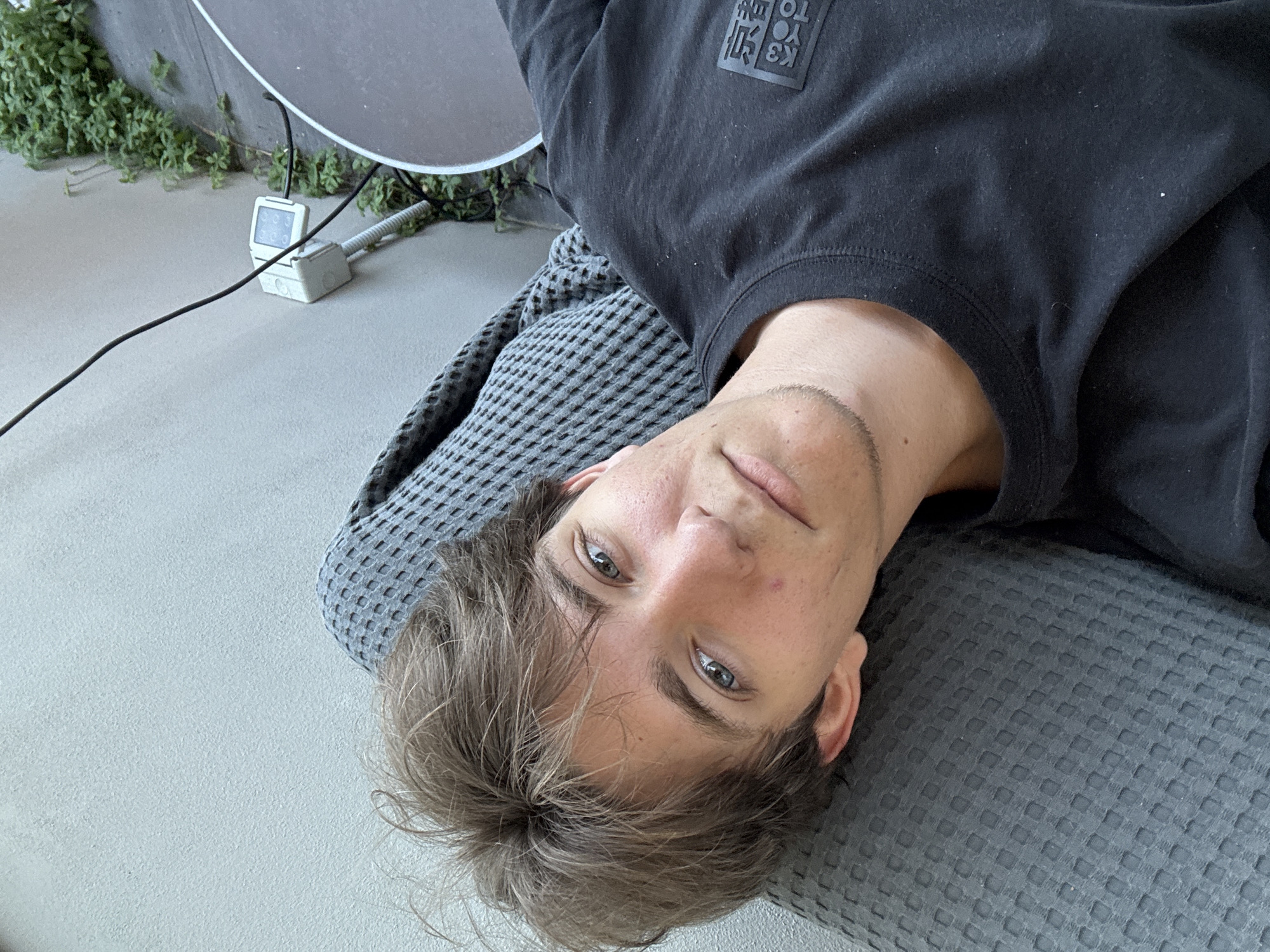 A young person with light brown hair and blue eyes is lying on a textured gray surface, possibly a sofa or cushion, with a gray wall in the background. They are wearing a black t-shirt with a black square logo containing Japanese characters. A black cable runs diagonally across the image from top left to the middle right, connected to a white electrical box mounted on the wall. Green vines are visible growing on the wall to the right of the electrical box. A portion of a large, round, dark gray object, possibly a table, is also visible on the right side of the frame. The scene appears to be outdoors, possibly in a courtyard or patio area, in the city of Cupra Marittima, Italy.