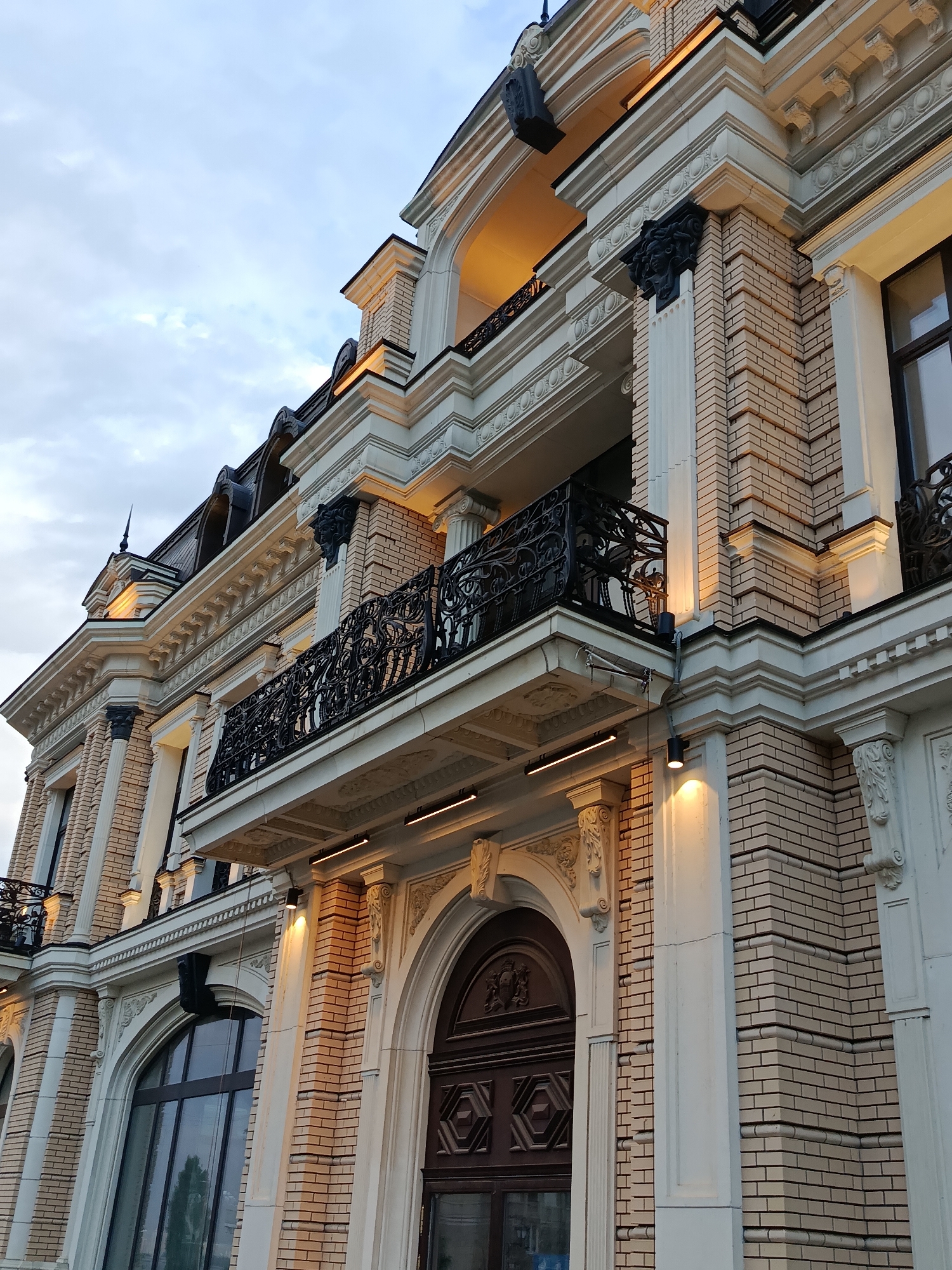 The image shows a low-angle view of a light-colored brick building with ornate architectural details, suggesting a grand or historical facade. The building has a balcony with intricate black wrought-iron railings, supported by decorative corbels. Above the balcony, there are columns and decorative cornices. The building is illuminated by warm, artificial lights, highlighting the textures of the brickwork and the architectural elements. The sky above is overcast with a mix of light and dark clouds. The overall impression is of a well-maintained, possibly upscale building. This building is located in the city of Saratov, Russia.