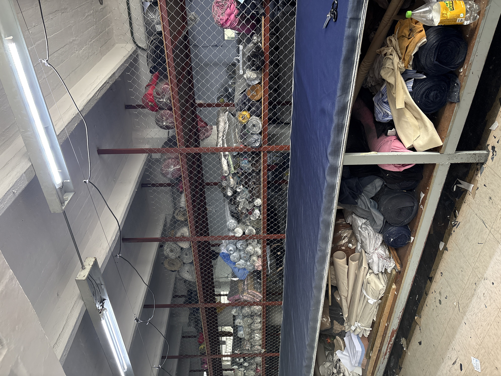 The image depicts a cluttered storage space in Mexico City, Mexico. Rows of stacked fabric rolls, secured behind wire mesh fencing, dominate the left side of the frame. A narrow shelf runs along the right side, crammed with rolls of fabric, loose clothing, a plastic bottle of yellow liquid, and miscellaneous debris. The space is lit by fluorescent tube lights mounted on the ceiling. The floor appears to be dirty and damaged. There are no people visible.