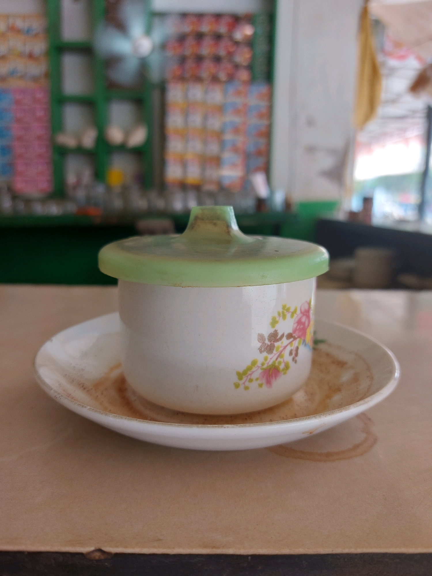 A close-up, eye-level shot shows a white ceramic cup with a pale green lid, resting on a white saucer. The cup is decorated with a floral pattern featuring pink flowers and green leaves. The saucer has visible stains, likely from a beverage. The scene appears to be set on a light brown, textured surface, possibly a table. In the blurred background, a green shelf unit is visible, stocked with various items, and a fan is mounted on the wall. The setting suggests a shop or eatery in Sumuragung, Indonesia.