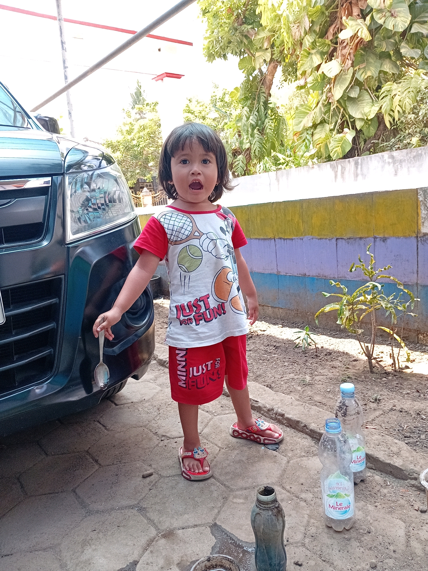 A young child, estimated to be between 2 and 4 years old, stands in front of a dark-colored vehicle. The child is wearing a red and white t-shirt with a graphic print and red shorts. On their feet are red sandals with cartoon character embellishments. The child holds a metal spoon in their right hand, which is resting on the front bumper of the car. The child's mouth is open, as if speaking or surprised.
The scene appears to be an outdoor setting, possibly a driveway or yard. To the left of the child is the front of the dark vehicle. A wall with horizontal bands of yellow, purple, and blue is visible behind the child. In front of the wall, a small plant with green and yellow leaves is growing. On the ground to the right of the child are two empty plastic water bottles, one of which has a blue cap and a label that reads "Le Minerale." Another plastic bottle, which appears to be a different brand and is partially filled with liquid, is also present on the ground. The ground surface is a mix of paved stone and dirt. Trees with lush green foliage are visible in the background. The lighting suggests it is daytime, with bright sunlight casting shadows.
The overall impression is a candid snapshot of a child engaged in an activity outdoors, with elements of everyday life in the vicinity of a residence in Gebang, Indonesia.