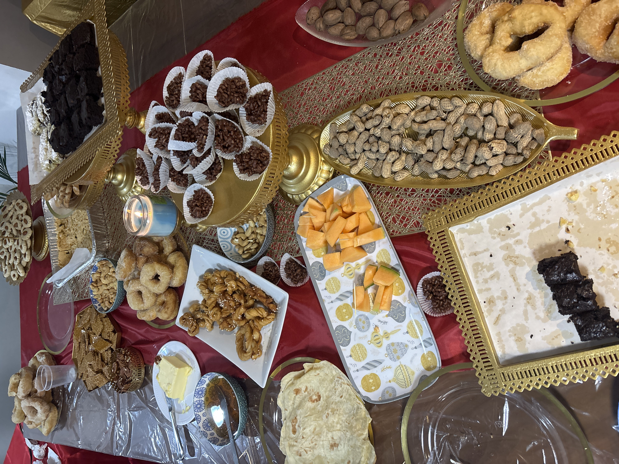 A high-angle shot shows a table laden with a variety of foods. The table is covered with a red cloth, partially obscured by a gold-patterned runner. On the left, a gold stand holds brownies and powdered pastries. Next to it are small paper cups filled with chocolate rice cereal treats. Further right are bowls of nuts, fried dough pastries, melon slices, and other unidentified desserts. A bowl of butter and a bowl of syrup are also present. The background is indistinct. The location is Huixquilucan de Degollado, Mexico. There are no people visible.