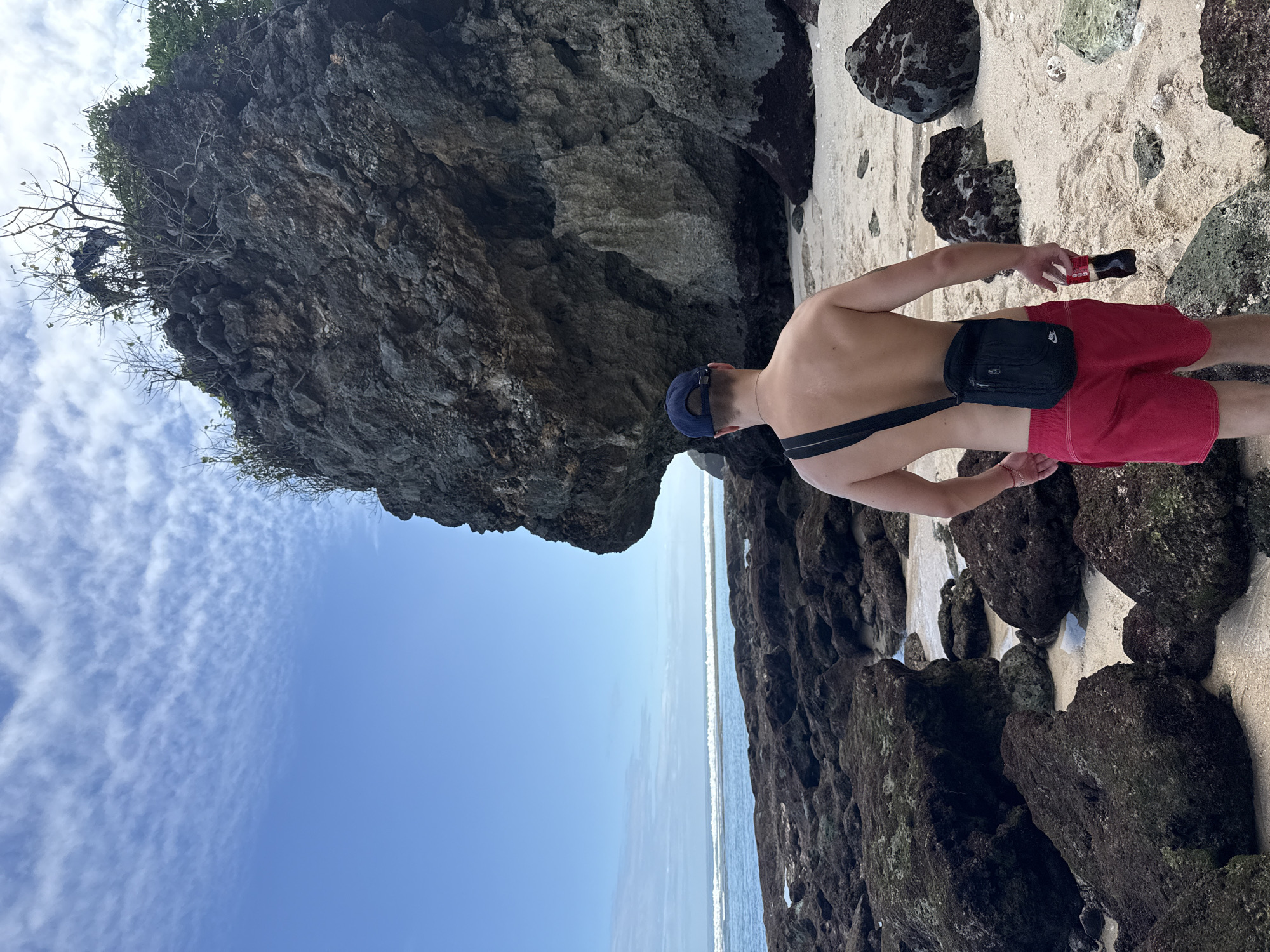 A shirtless man wearing red shorts, a blue baseball cap, and a black fanny pack stands on a sandy beach beside large, dark rocks. He is holding a dark object in his right hand. The beach appears to be strewn with similar dark rocks and patches of sand. Beyond the rocks, the ocean meets a clear blue sky with wispy clouds. A large, gray cliff looms over the man's left side, with vegetation growing on its top.
The location is Kuta Selatan, Indonesia.