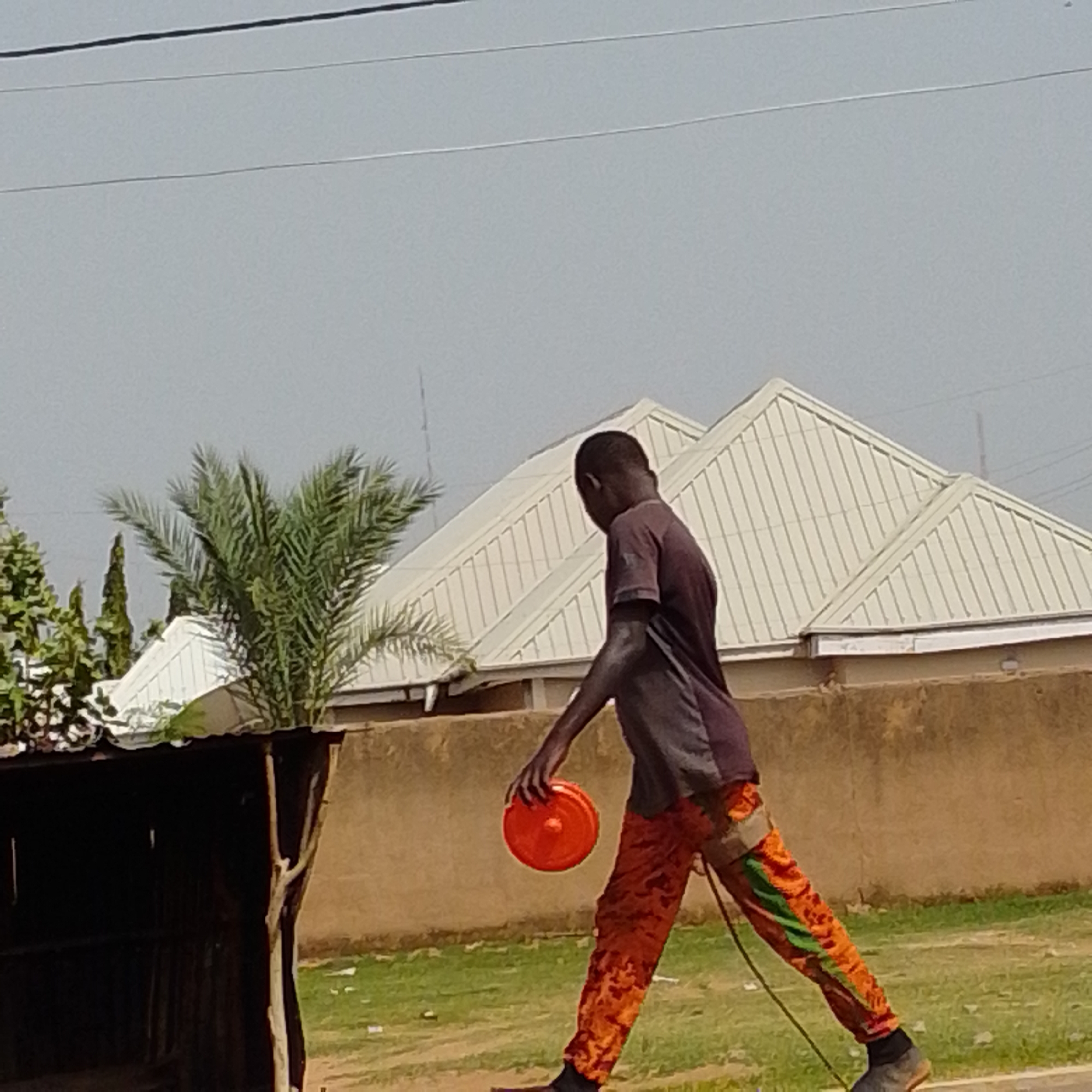A young Black male is walking outdoors, holding a round, orange object in his right hand. He is wearing a dark gray t-shirt and orange pants with a patterned design. He is walking on a paved surface, and a grassy area with some white litter is visible behind him. In the background, there is a long, light-colored building with a corrugated metal roof. To the left of the building, a palm tree and other greenery are visible. The sky is overcast. The location is identified as Tempure, Nigeria.