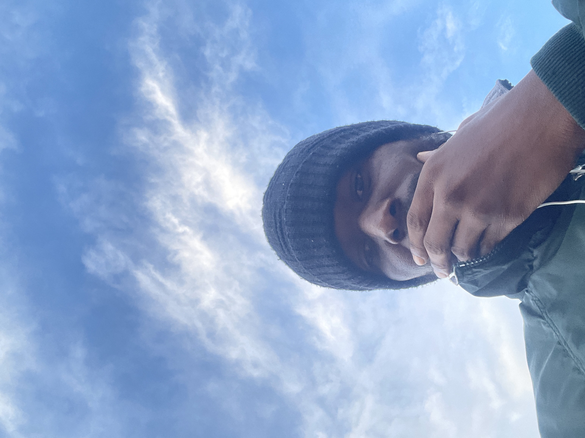 A low-angle, close-up, outdoor, selfie-style photograph shows the upper body and head of a dark-skinned person wearing a dark knitted beanie and a dark green jacket. The person's right hand is raised to their mouth, partially obscuring it, and their left ear is visible with a white earbud wire. The background consists of a bright blue sky with scattered white clouds. The person appears to be looking directly at the camera. The image does not provide any specific indicators of its location within Abuja, Nigeria.
