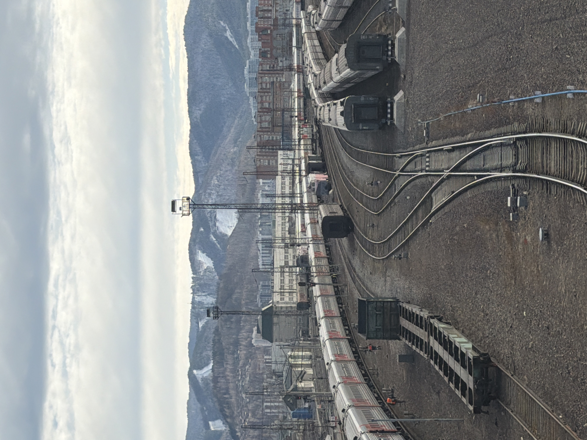 The image depicts a train yard in Krasnoyarsk, Russia. Several trains are on multiple tracks with switches allowing movement between lines. Mountains rise in the background with some snow on their slopes. The weather appears overcast, with a cloudy sky. There are industrial buildings and infrastructure surrounding the train yard including what looks to be a train operator's tower. There are a few visible figures wearing orange vests near the trains, possibly railroad employees.