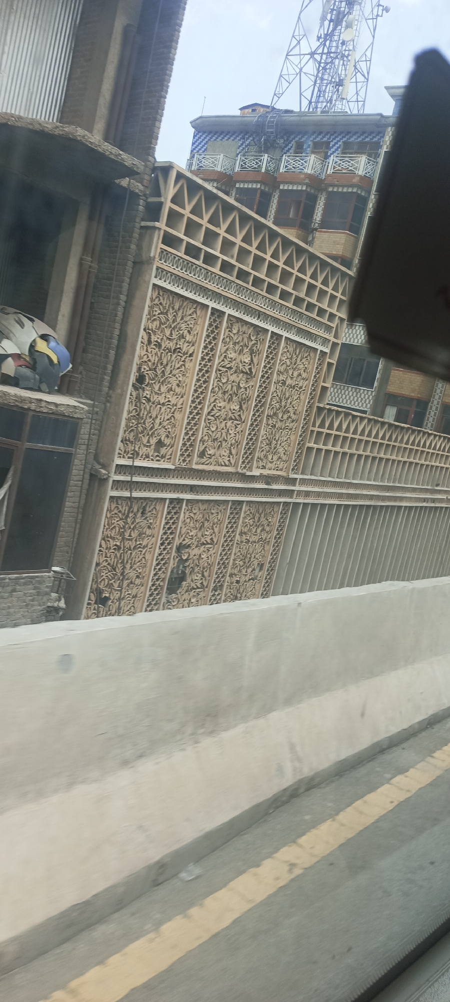The image shows a detailed view of the facade of a building with intricate latticework and decorative carvings. The building features multiple levels with balconies and windows. A prominent element is the ornate screen-like structure with repeating floral patterns, suggesting a traditional architectural style. Above this, there are sections with a grid-like pattern and some areas with blue and white patterned tiles. A large communication tower is visible on the rooftop of a building in the background. In the foreground, there is a concrete barrier and a yellow line marking on a road, indicating that the photo was likely taken from a moving vehicle. The environment appears to be urban, with other buildings and structures visible in the distance. The lighting suggests it is daytime. The image is oriented sideways, with the top of the buildings appearing on the left side of the frame. There are no people clearly visible in the image.