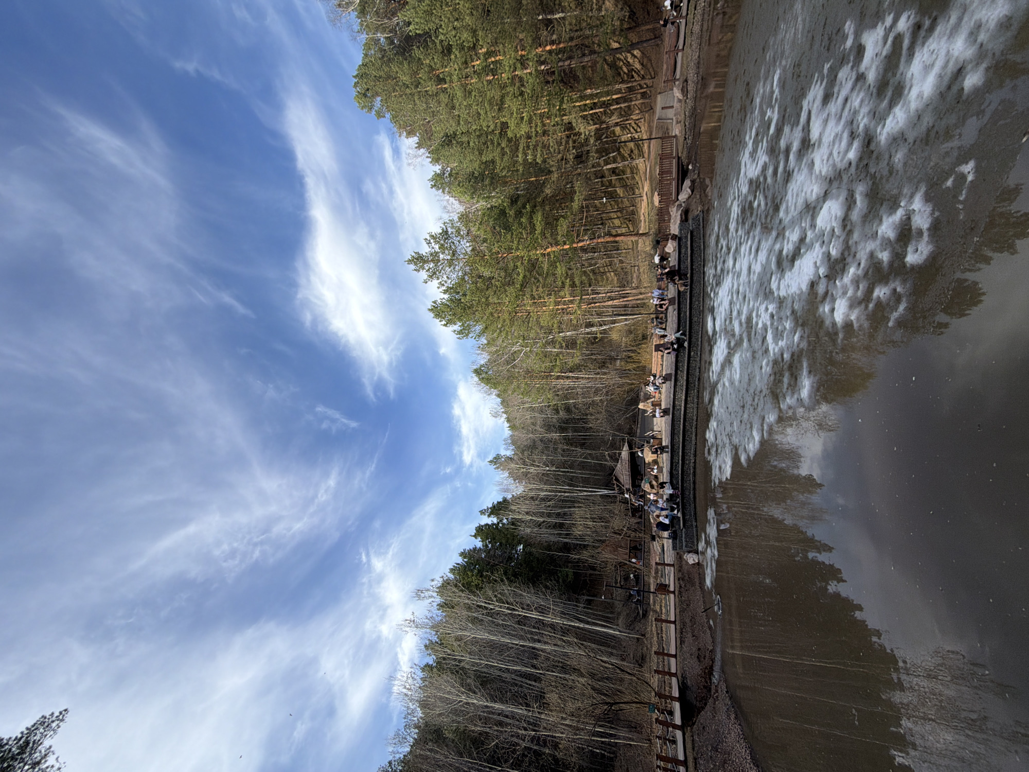 The image captures a serene scene in Krasnoyarsk, Russia. A line of people sit along a wooden boardwalk bordering a body of water. The water reflects the sky and surrounding trees, with patches of ice or snow present on the surface. Dense forest lines the opposite side of the boardwalk. The sky is blue with wispy clouds. It is a sunny day.
