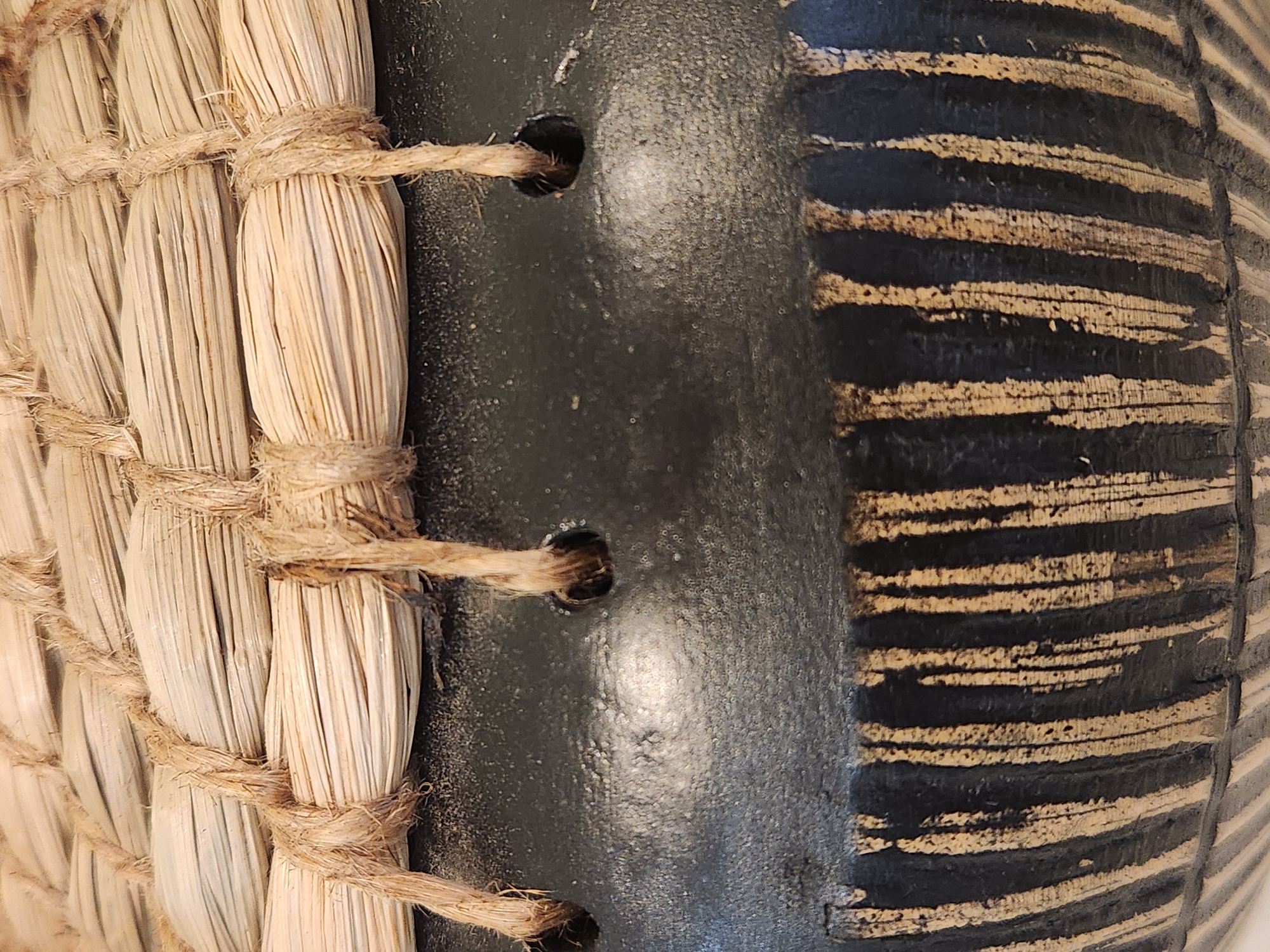 The image is a close-up of a woven straw lid with natural fiber rope lashing, placed on a dark, textured surface. The surface appears to be ceramic or painted metal and has vertical stripes of light brown and dark brown/black. Two visible holes pierce the dark surface, through which the rope lashing passes. The scene is lit, highlighting the textures of the straw, rope, and the patterned surface. No specific location in Maitland, United States of America, can be determined from the visual information provided in the image.