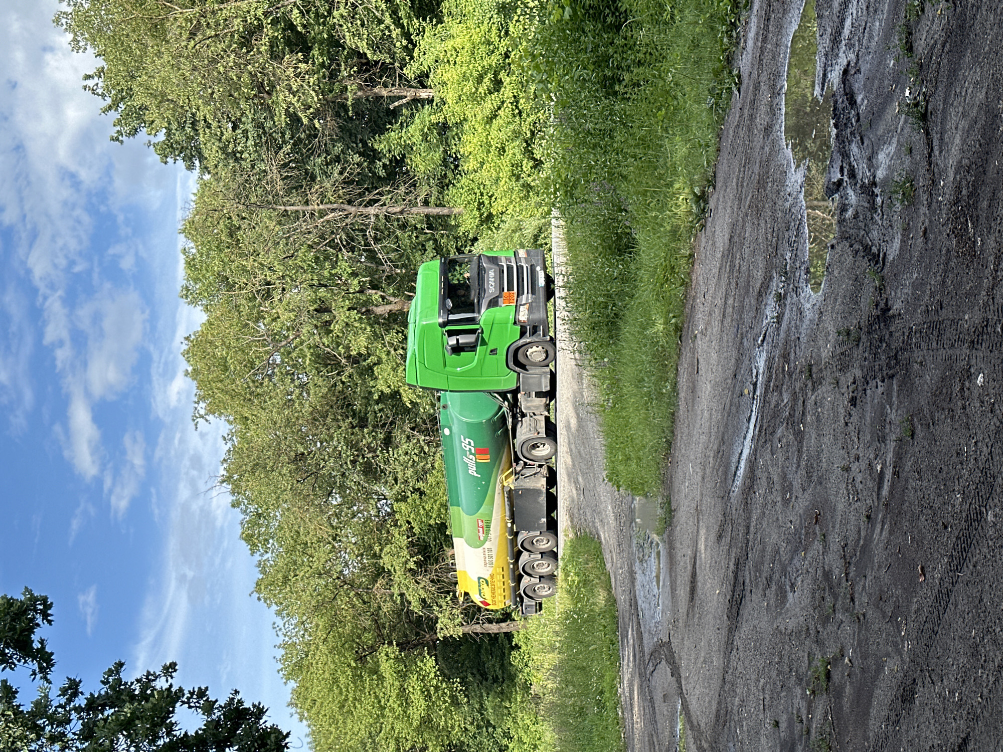 In Shatava, Ukraine, a bright green Scania tanker truck with a yellow and green striped tank is driving on a muddy, unpaved road. The truck is carrying what appears to be fuel, as indicated by the "pulls 95" labeling on the tank. The road is wet and has tire tracks, suggesting recent rain. The truck is surrounded by lush green trees and foliage, typical of a rural or wooded area. The sky is blue with scattered white clouds.