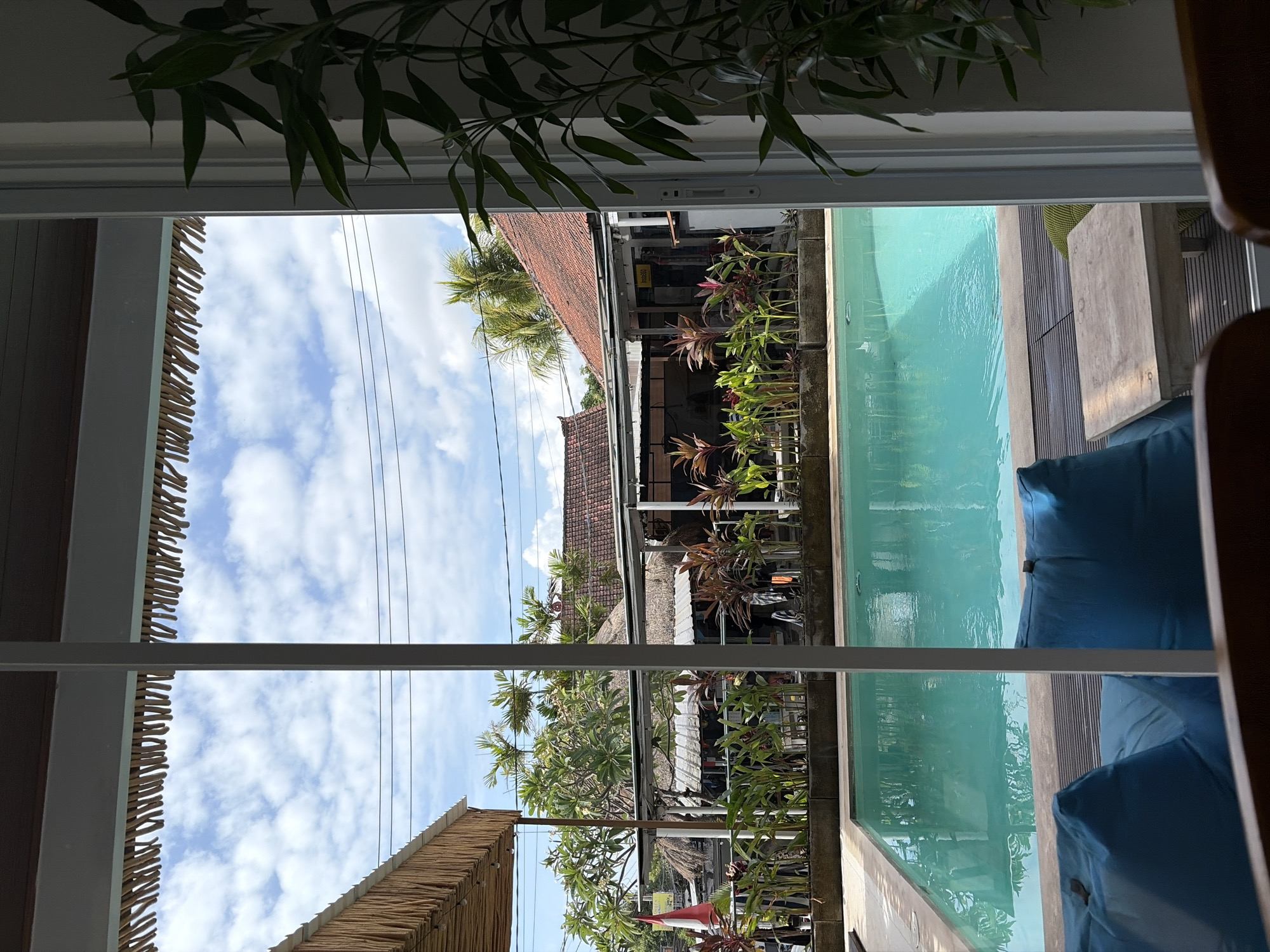 The image captures a view of a swimming pool and surrounding area through a large glass door. The swimming pool, filled with clear turquoise water, is the central element. Along the edge of the pool is a deck made of dark wood planks. In the foreground, two large blue beanbag chairs are visible, along with a small wooden table with a light-colored top and dark legs.
Beyond the pool, there is a strip of garden with various plants, including tall green stalks and plants with reddish-purple leaves. Behind the garden, there are several structures with thatched roofs and red tiled roofs, suggesting a tropical or resort-like setting. Trees with green foliage are scattered throughout the scene. The sky above is partly cloudy, with a mix of blue sky and white clouds. The overall impression is a relaxed outdoor environment, likely in a warm climate.
The location is identified as Kubu, Indonesia. The image appears to be taken from an indoor space, looking out towards the pool and courtyard.