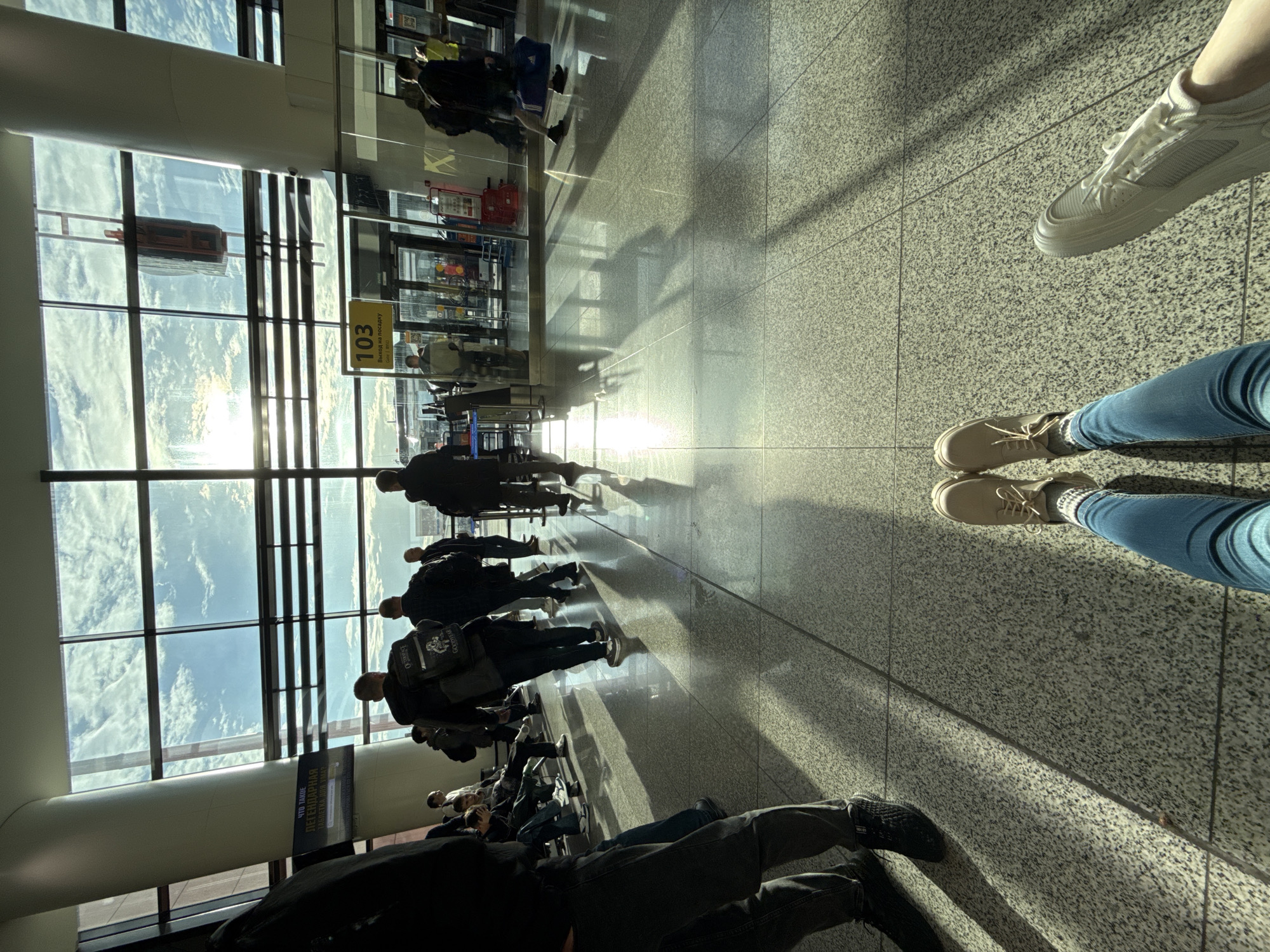 The image depicts the interior of an airport terminal in Khimki, Russia. Sunlight streams through large windows, casting long shadows across the tiled floor. A queue of people are waiting near gate 103. Two pairs of legs and feet are visible in the foreground; one with light-colored sneakers and blue jeans, and the other wearing beige shoes with blue jeans and patterned socks.