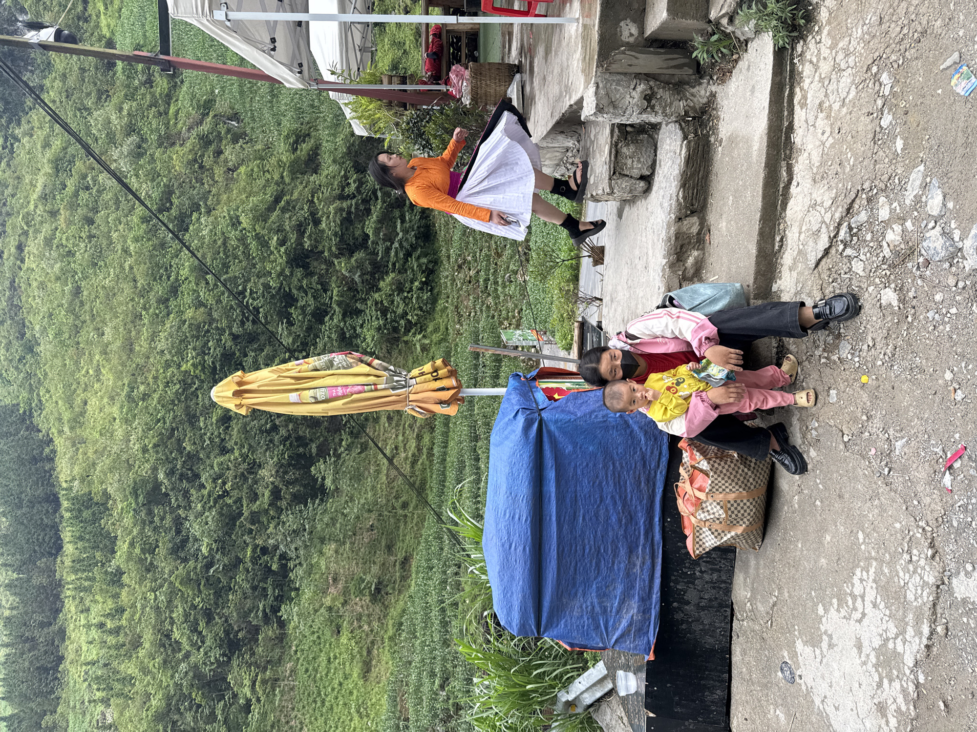 The image shows a scene in Yên Minh Commune, Vietnam. A woman in an orange shirt and white skirt stands on a concrete step, seemingly posing or dancing. Below her, another woman sits on the ground holding a baby. A yellow umbrella and blue tarp are nearby, partially obscuring the background of lush green hills and vegetation. A large checkered bag is positioned near the seated woman. The ground is concrete and appears worn. The scene suggests a roadside stop or informal market area.