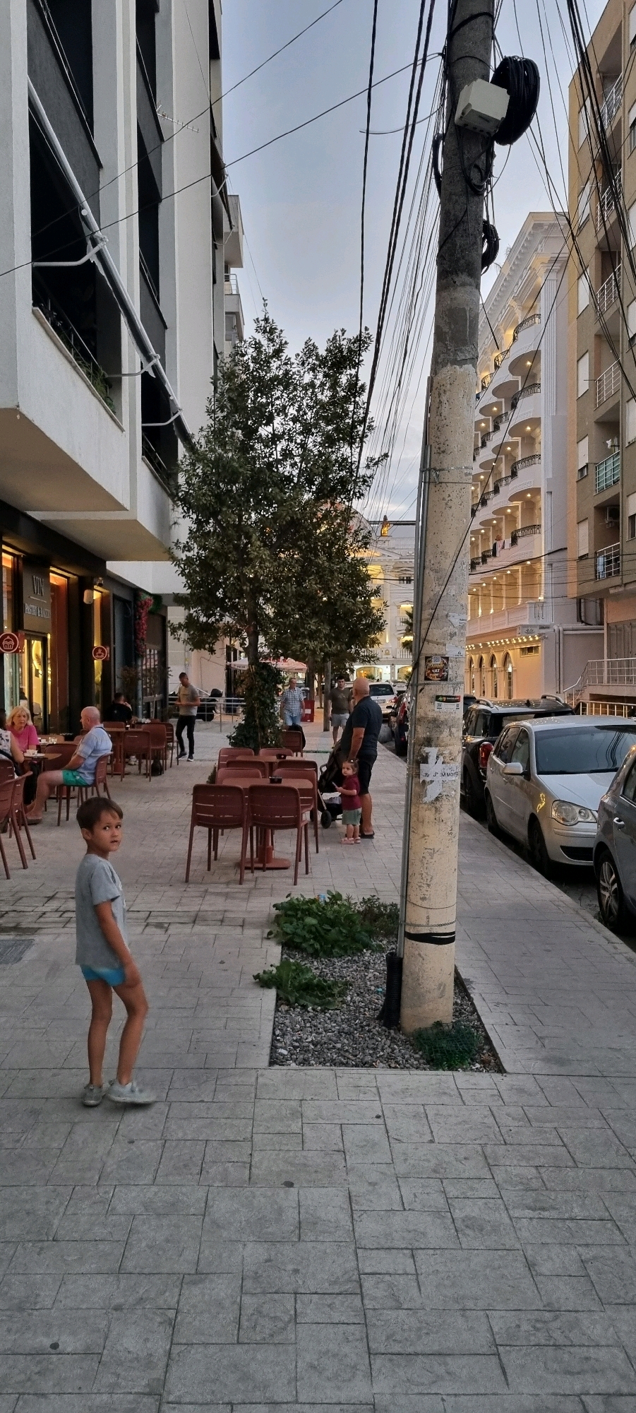 This image depicts a street scene in Bashkia Durrës, Albania. A young boy in a grey t-shirt and blue shorts stands on a paved sidewalk, looking towards the camera. In the background, an outdoor seating area for a restaurant is visible, with tables and chairs. Several people are seated at the tables. To the right of the seating area, a utility pole with numerous wires is prominent, along with a parked silver car. Further down the street, other buildings and parked vehicles can be seen. The sky is overcast, suggesting it might be late afternoon or early evening.