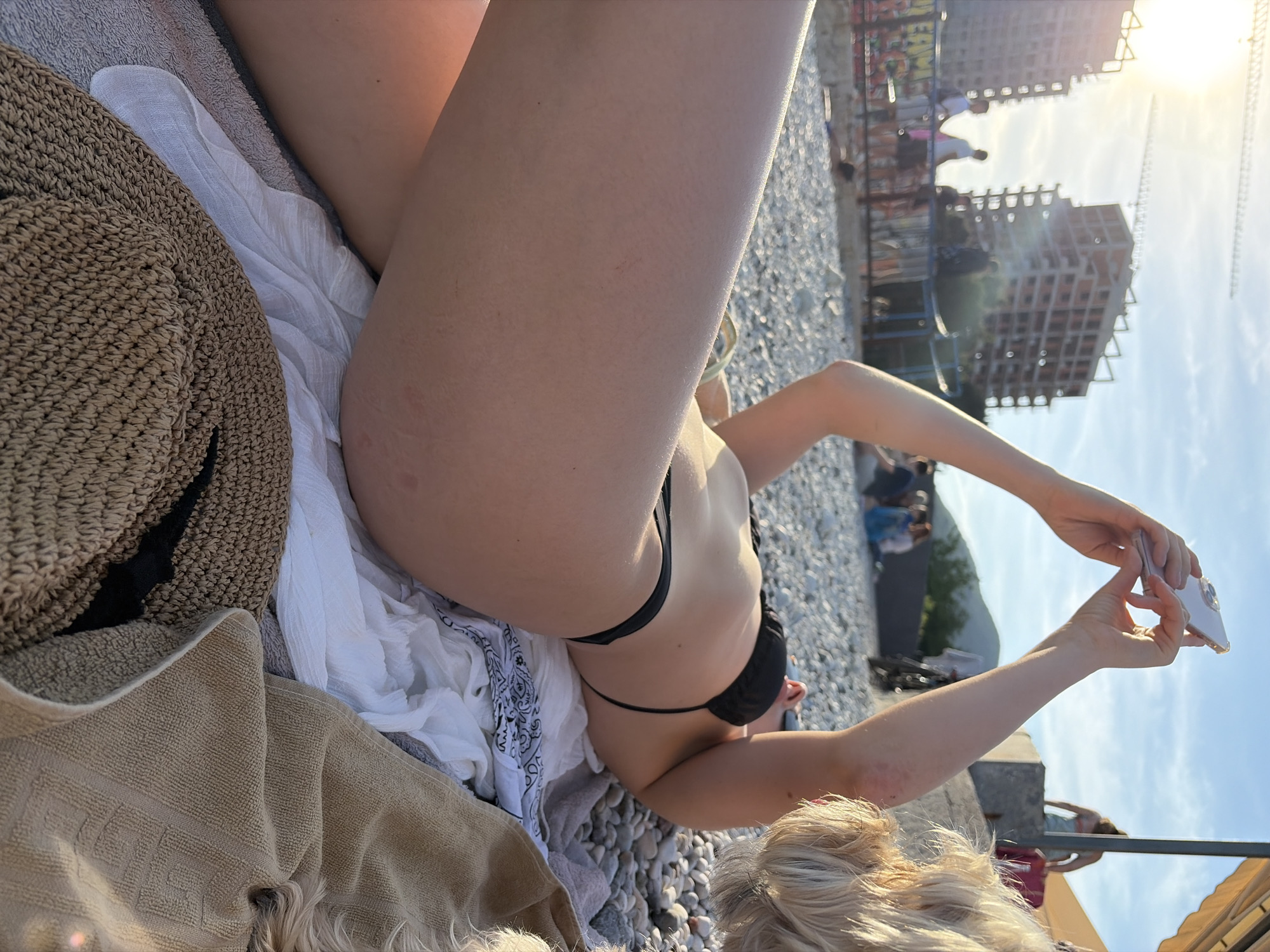 A woman in a black bikini is lying on a beach in Budva, Montenegro. She is using her smartphone to take a picture. A brown towel and hat are near her, while some of her body is lying on a white scarf. People are scattered throughout the background near a newly constructed building with cranes. The sky is sunny and blue.