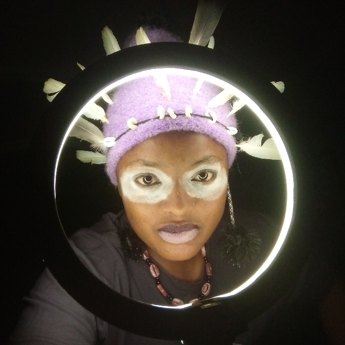 A person with dark skin, adorned with white face paint around the eyes and lips, wears a lavender knit cap with white feathers and shells. A black cord encircles the cap. She sports a beaded necklace and a dangling earring. The person is positioned within a circular light ring, creating a stark contrast against the black background.