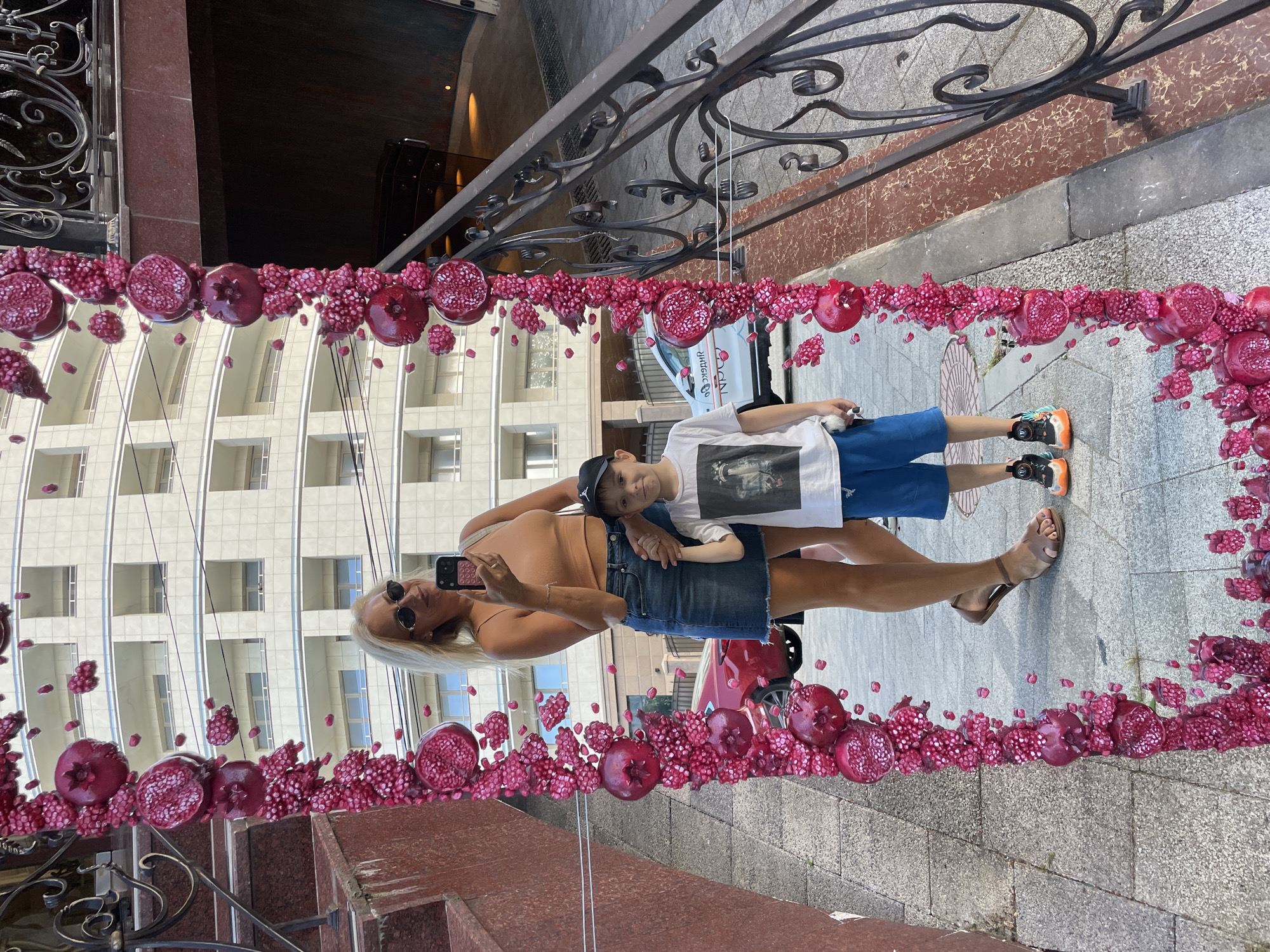 In Sochi, Russia, a woman with blonde hair and sunglasses is taking a selfie in a large, ornate mirror decorated with pomegranates. She is wearing a tan tank top and a denim skirt. Beside her, reflected in the mirror, is a young boy wearing a black baseball cap, a white t-shirt with a graphic print, and blue shorts. He is holding the woman's hand. The background shows a modern, multi-story building and a street scene with a red car and a white car parked. The pavement is made of grey tiles, and a decorative metal railing is visible to the right of the mirror.