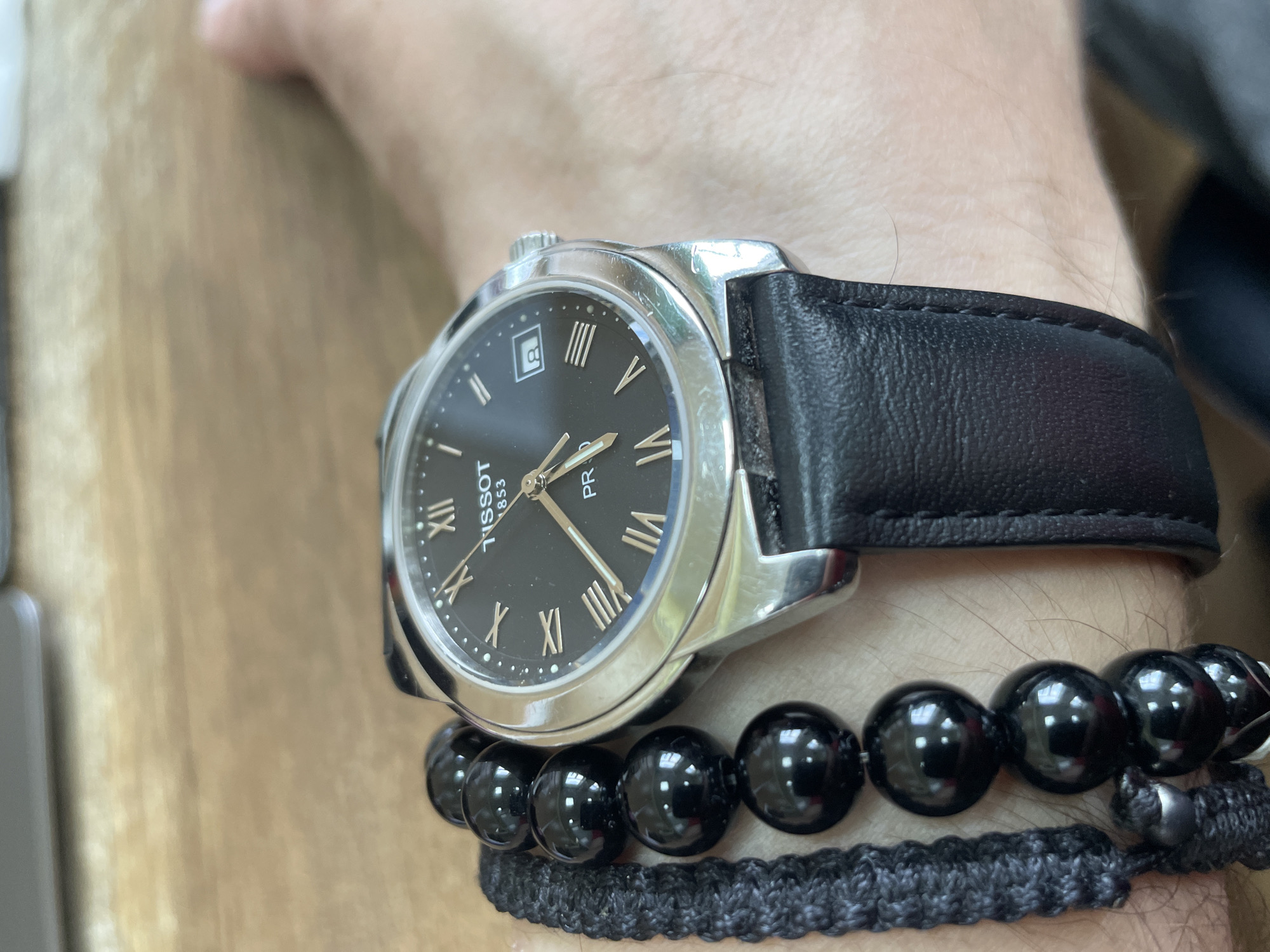 The media file shows a close-up view of a person's wrist wearing a Tissot PR 100 watch and two bracelets. The watch has a black dial with Roman numeral hour markers and a date window at the 3 o'clock position. The watch band is made of black leather. Below the watch, there are two bracelets: one made of black, polished beads, and another woven, dark gray bracelet. The background appears to be a wooden surface, possibly a table or desk. The lighting suggests it is daytime, indoors, with no clear indication of the specific time. The setting is casual, possibly a home or office environment. The image is focused on the accessories worn on the wrist, highlighting the watch and bracelets. There is no explicit information about the person, emotions, weather, or specific location beyond the provided context of Moscow, Russia. Visible text on the watch includes "TISSOT 1853" and "PR 100".
