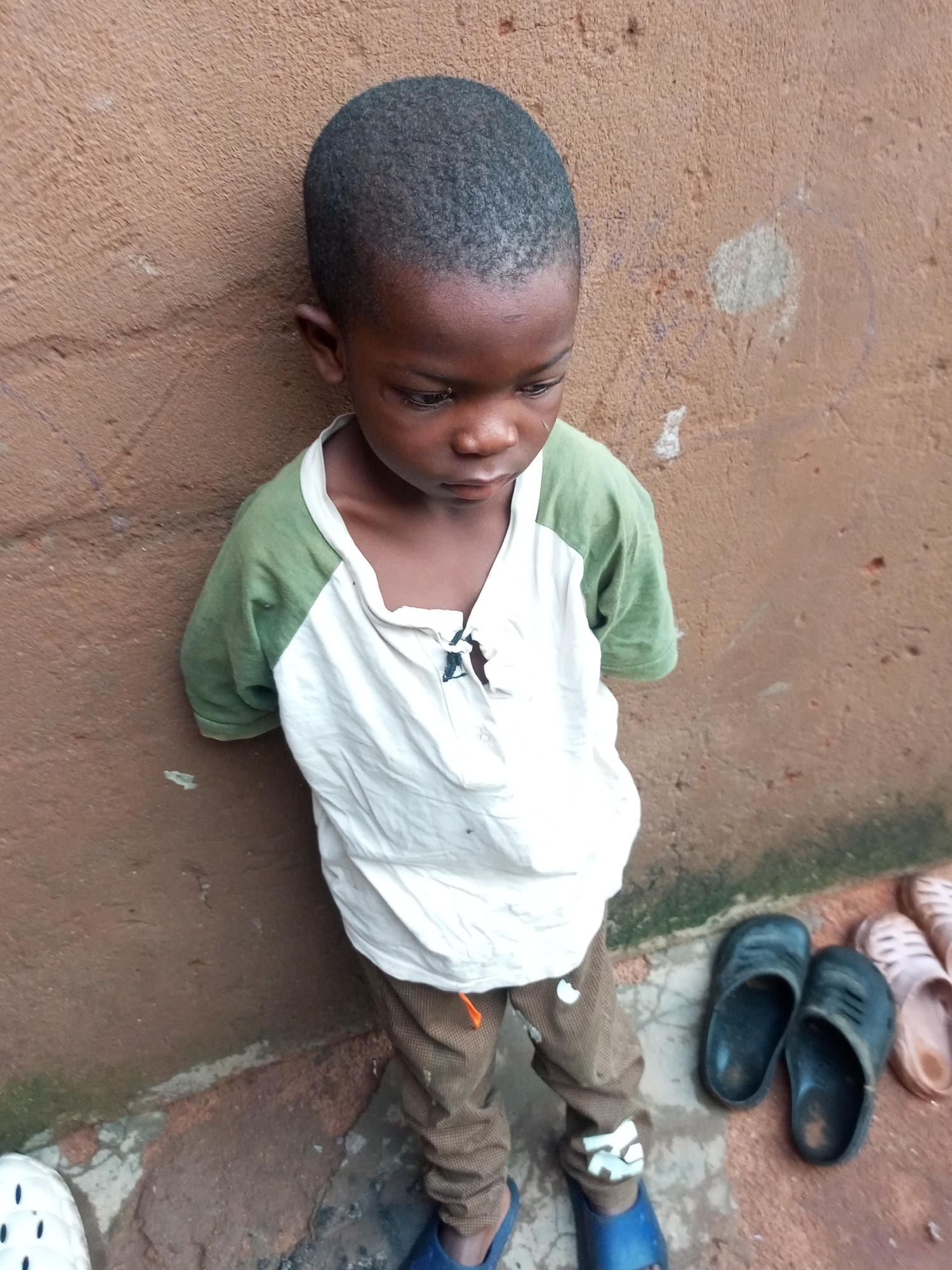 A young child is standing in front of a brown, textured wall in Tempure, Nigeria. The child, facing downwards and to the right, wears a white t-shirt with green sleeves and brown pants. They have on blue sandals. To the right of the child, there are several pairs of sandals on the ground.