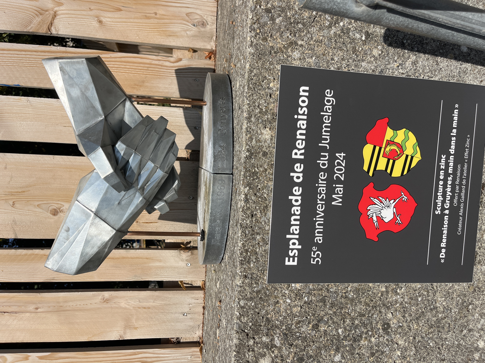 The image depicts a modern metal sculpture of two hands clasped together, positioned in front of a wooden fence. The sculpture is mounted on a circular base with inscriptions that appear to be "Renaison 1969" and "2024 Gruyères".
In the foreground, a dark gray sign with white text provides information about the artwork. The sign reads: "Esplanade de Renaîson 55e anniversaire du Jumelage Mai 2024". Below this, there are two coats of arms. The left coat of arms is red with a white bird. The right coat of arms is divided vertically, with the left half yellow and black striped, and the right half red and green with a crest in the center. Further text on the sign reads: "Sculpture en zinc « De Renaîson à Gruyères, main dans la main » Offert par Renaîson Créateur Alexis Gaillard de l'atelier « Effet Zinc »".
The setting appears to be outdoors, with natural light casting shadows. The ground is a rough, textured concrete. The overall impression is that of an artistic installation commemorating a twinning or anniversary event. Based on the text on the sign mentioning "Gruyères", the location is likely in or near Gruyères, Switzerland, although the sign specifically mentions "Esplanade de Renaîson", suggesting a specific area named after Renaîson. Epagny, Switzerland, is not explicitly mentioned on the sign.