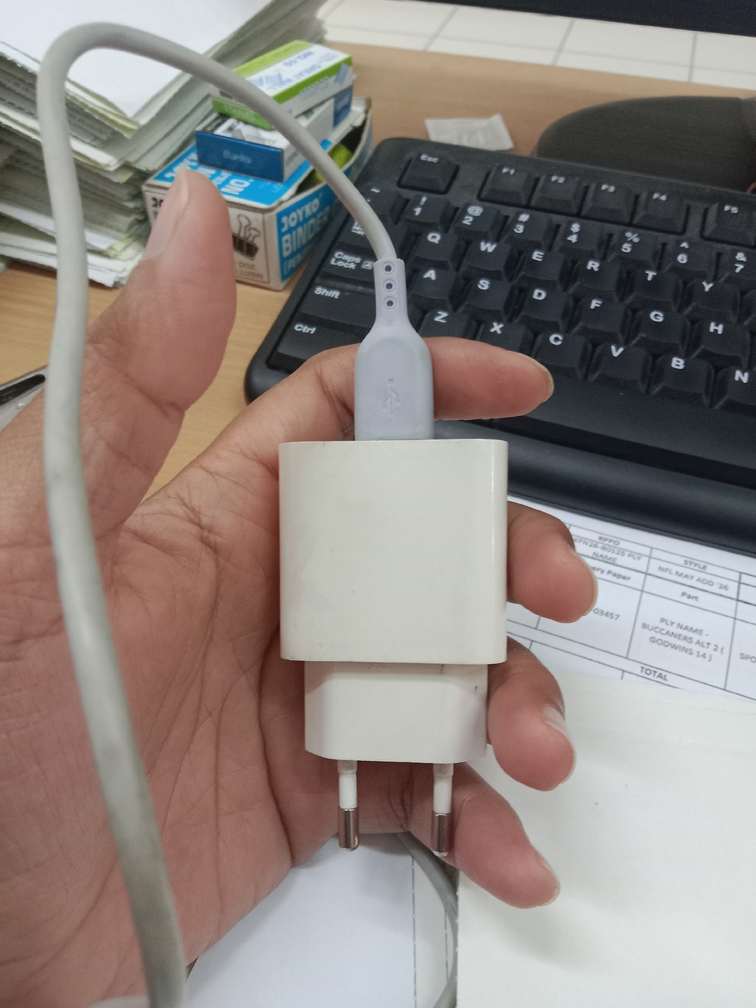 The image shows a close-up of a hand holding a white power adapter with a USB cable plugged into it. The hand is positioned in front of a black keyboard, and in the background, there are stacks of paper and office supplies, including boxes of "JOYKO BINDER CLIPS." The overall setting appears to be an office environment. There is no information in the image to suggest a specific location within the city of Semarang, Indonesia.
