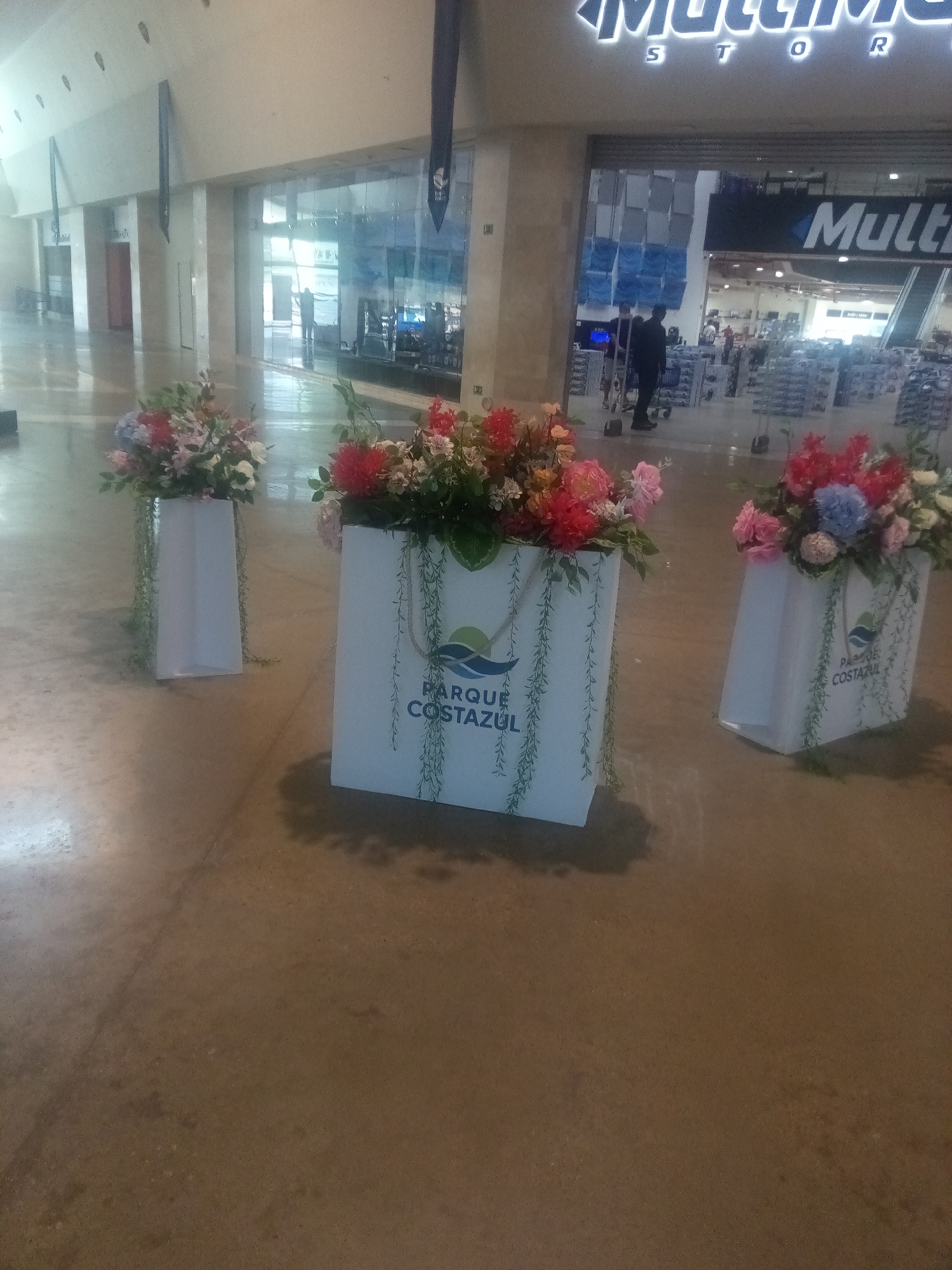 In the interior of a brightly lit shopping mall in Pampatar, Venezuela, three large white decorative planters are arranged. The planters are adorned with green vine-like drawings. The planter in the center and the one on the right display a logo with blue waves and the text "PARQUE COSTA ZUL". All three planters are filled with vibrant floral arrangements featuring red, pink, blue, and white flowers, as well as green foliage. In the background, a store with a large sign that reads "Multi" is visible through a glass storefront. Several people are walking in the background, some are seen pushing shopping carts. The floor of the mall is a polished, light-colored surface.