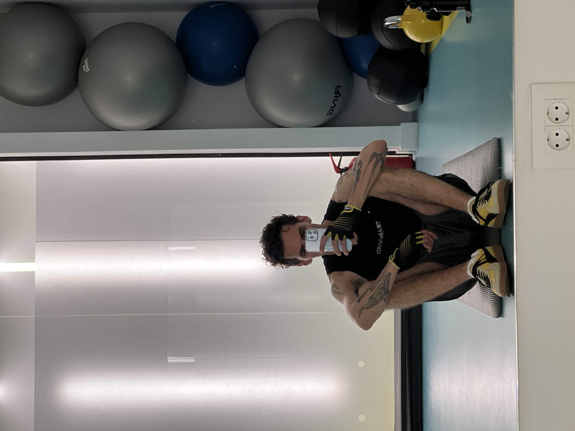 A man is sitting on a gray striped mat in what appears to be a gym setting. He is wearing a black T-shirt with white lettering, dark shorts, yellow and black sneakers, and fingerless gloves. He is holding a white smartphone in front of his face, presumably taking a selfie. His legs are crossed, and his arms are resting on them. Tattoos are visible on his forearms.
Behind him, there is a mirrored wall reflecting the scene. Above the mirrored wall, a shelf holds several exercise balls of various colors, including gray, blue, black, and yellow. To the right of the man, a light blue wall is visible, with a white electrical outlet mounted on the wall.
It is not possible to confirm the location in Boreti, Montenegro from the information available in this image.