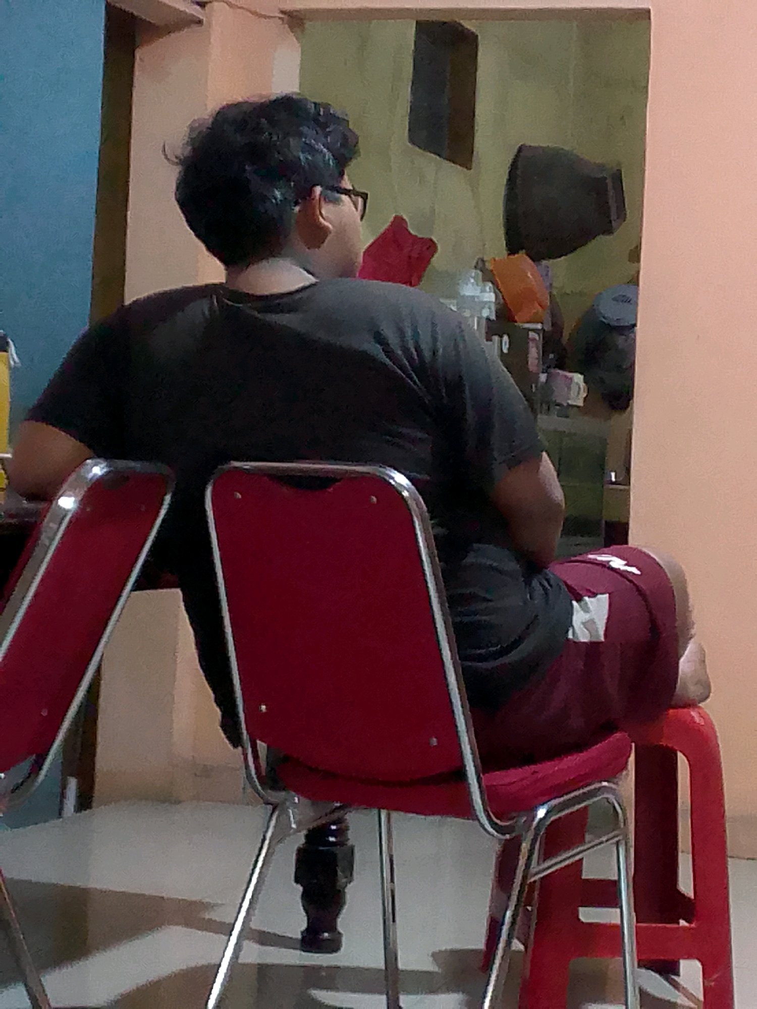 A person is seated in a red plastic chair with their back to the camera. They are wearing a black t-shirt and dark red shorts with a white emblem on the left leg. The person has dark, curly hair and appears to be wearing glasses. The chair is situated indoors, likely in a residential setting. In the background, there are other chairs, shelves with various objects, and a wall painted in muted tones. The lighting suggests it may be evening or indoors with artificial light. The environment appears somewhat cluttered. The image does not provide specific indicators of the city of Gunungmasigit, Indonesia.