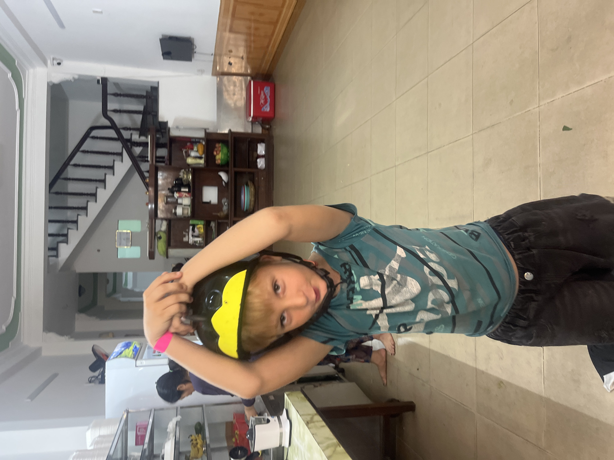 A young boy is standing in a tiled room, wearing a teal shirt with a graphic print and dark shorts. He has a black and yellow helmet on his head and a pink wristband on his left arm. His hands are on top of the helmet, holding it in place. In the background, a staircase with a wooden railing is visible, as is a wooden cabinet filled with various items, including bottles and bowls of fruit. A red cooler is on the floor to the right of the cabinet. There are shelves with kitchen appliances, including a white electric kettle, and a person with dark hair in the background to the left. The overall environment suggests a domestic setting, possibly a kitchen or dining area, within a building in Da Nang, Vietnam.