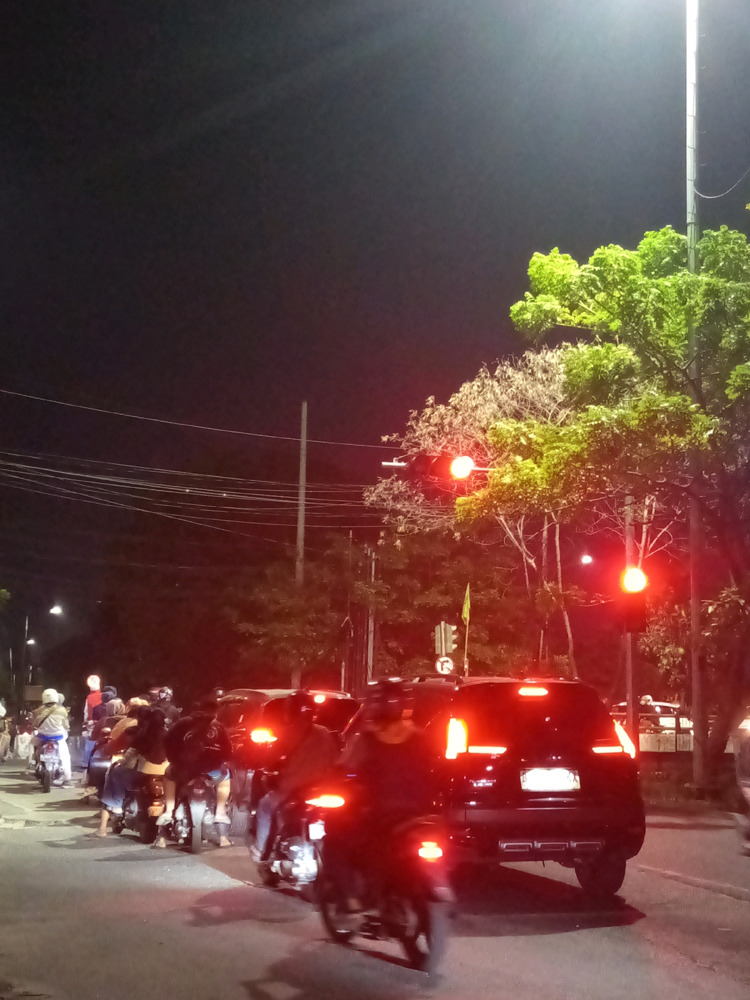 The image depicts a traffic scene at night in Bekasi, Indonesia, with several motorcycles and a black SUV stopped at a red traffic light. The sky is dark, illuminated by streetlights and the taillights of the vehicles. Lush green trees are visible on the right side of the frame, contrasting with the dark night. Several individuals are seated on motorcycles, with their bodies obscured by the low light and the angle of the shot. The traffic light signals a red light, indicating that vehicles are required to stop. The environment appears to be a street in an urban area, suggested by the presence of traffic signals, power lines, and vehicles.