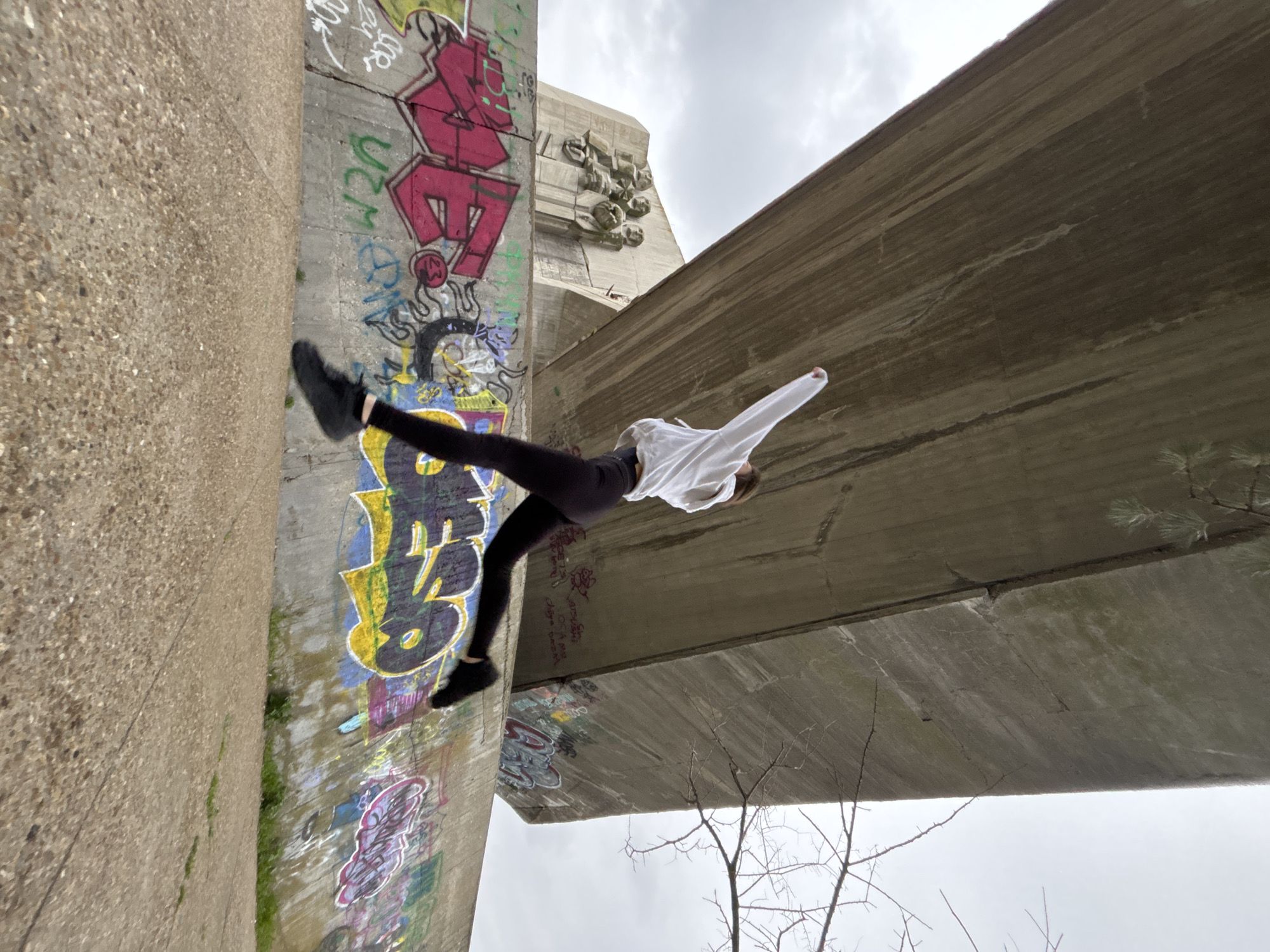 A person is captured mid-air, seemingly in motion between two concrete structures adorned with graffiti. The person is wearing dark leggings, black shoes, and a white top. One leg is extended upwards towards the left side of the frame, near a concrete wall covered with colorful graffiti. The other leg is bent downwards.
The backdrop consists of two large concrete structures, one of which is a wall heavily decorated with graffiti, and the other appears to be the underside of a bridge. The space between these structures allows a view of the overcast sky. A few bare branches of a tree are visible in the lower right corner of the image.