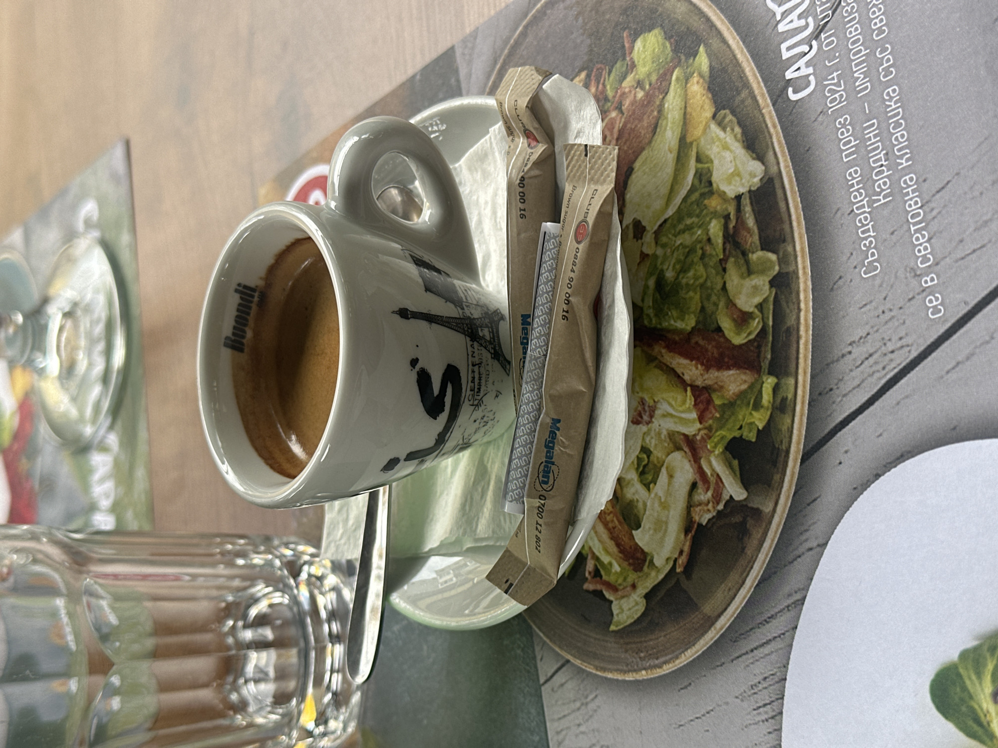 The image presents an overhead perspective of a table setting in an establishment located in Sofia, Bulgaria. In the left foreground, a clear, fluted glass contains water. Adjacent to it on the right, a white espresso cup, branded "Buondi" and featuring a graphic of the Eiffel Tower, holds an espresso and rests on a matching white saucer. A small silver spoon is positioned between the glass and the cup. On the saucer, a white paper napkin supports two brown sugar packets; one is labeled "Megalàn" with the Bulgarian phone number "0700 12 802," and the other is marked "CLUB Brown sugar" with the Bulgarian phone number "0884 90 00 16." All items are placed on a printed placemat designed to mimic light gray wooden planks. The placemat displays an image of a salad and partial Bulgarian text, which translates to "Created in 1924 by Ita... Cardini – improvisa... in world classic with fre..." (referencing the Caesar salad). A segment of a plain white plate with a single green leaf is visible in the bottom left corner. The background consists of a light wooden tabletop, with an out-of-focus area revealing another glass on a patterned green placemat. No individuals are depicted in the scene.