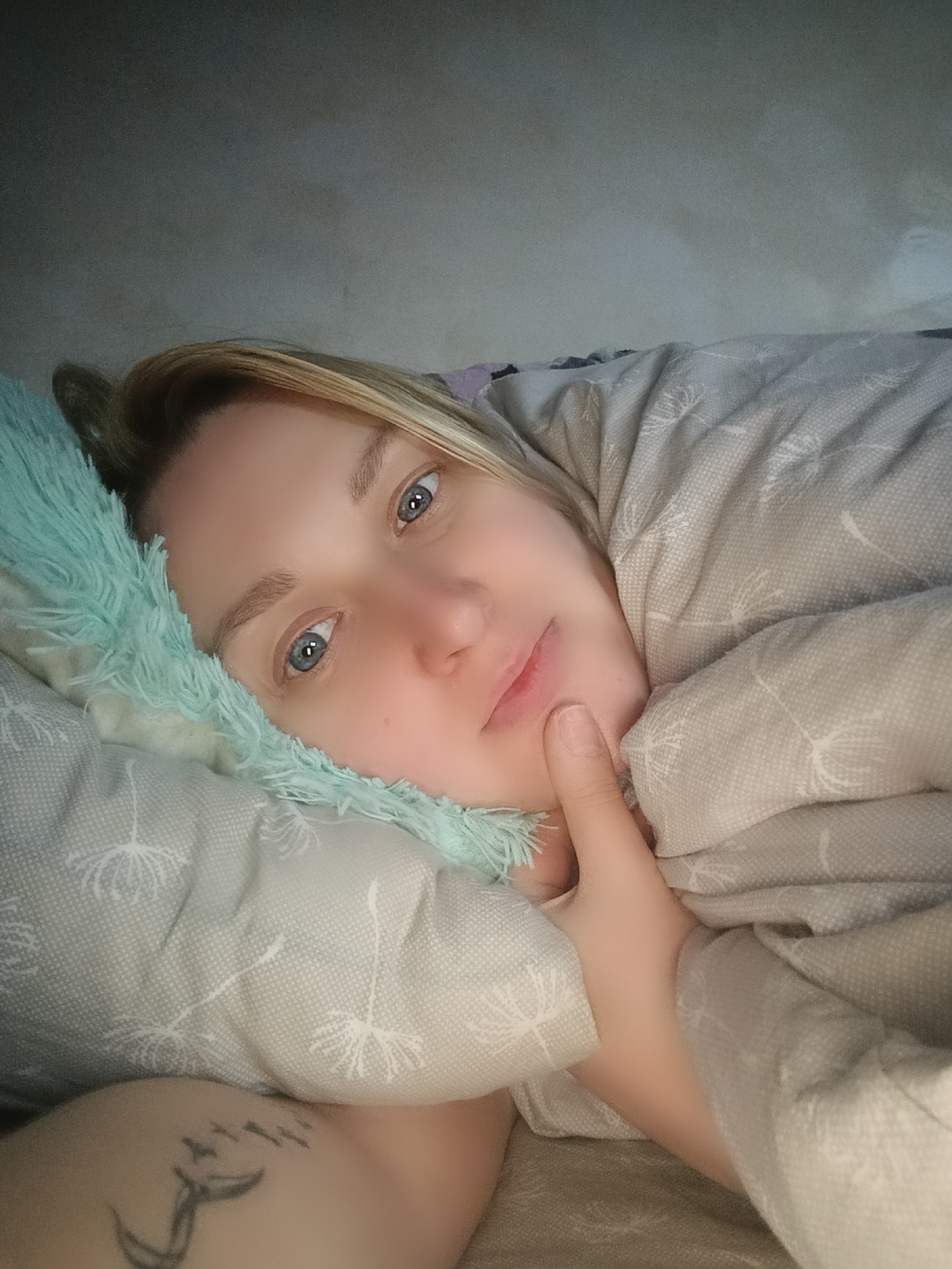 A fair-skinned woman with blonde hair and blue eyes is lying down in a bed. Her head rests on a pillow with a light grey floral pattern, overlaid with a fluffy turquoise pillow. She is partially covered by a grey patterned blanket, which is pulled up around her. Her right hand is touching her face near her mouth. A tattoo of birds in flight is visible on her left arm. The background is a slightly out-of-focus wall or ceiling. The image does not provide sufficient information to determine a location in the city of Chaikovsky, Russia.