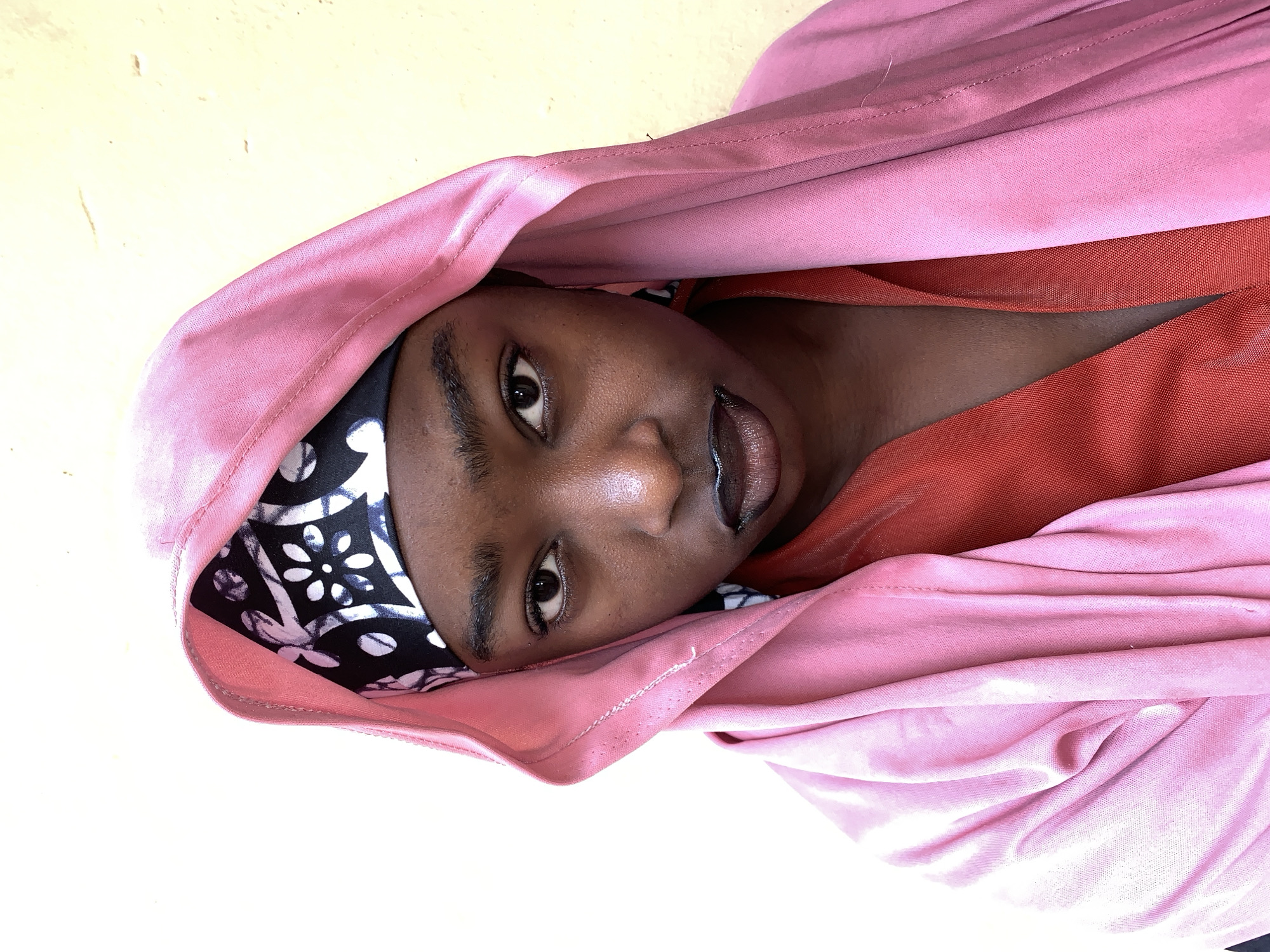 A close-up, portrait-style photograph shows a Black person with brown skin looking directly at the viewer with a serious expression. The person's head is covered with a headscarf with a black and white pattern and a light pink fabric draped over their head, creating a hooded effect. The person also wears a red garment. The photograph's background is a light yellow color. There is no indication of whether or not the image was taken in the city of Rafin Par, Nigeria.