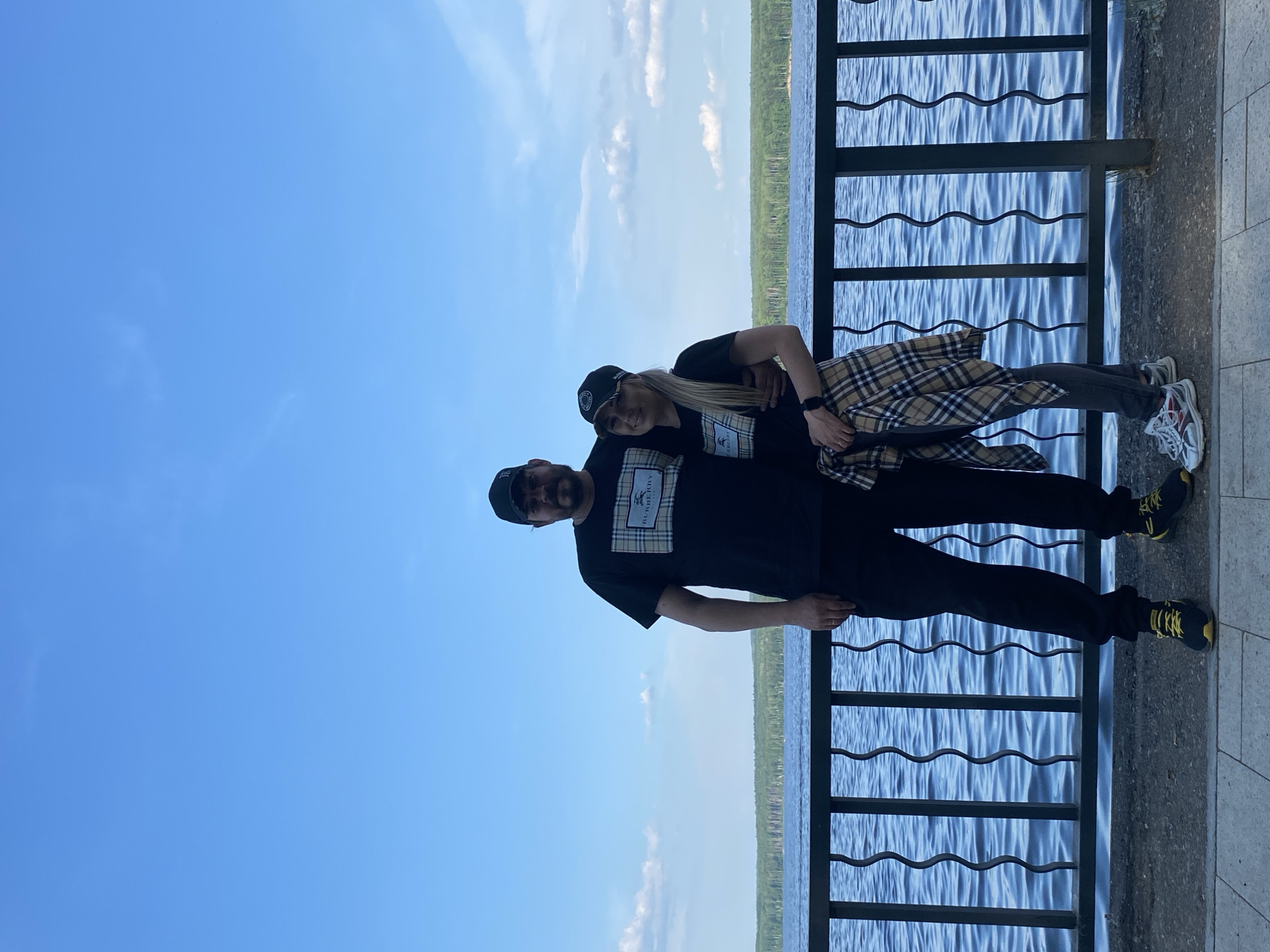 This full-frame, eye-level, vertical outdoor photograph captures a couple standing on a paved embankment in the city of Chaikovsky, Russia, with a large body of water and a treeline in the background. The sky above is clear blue with scattered white clouds. A dark metal railing with a wavy design runs horizontally across the lower portion of the frame, separating the embankment from the water.
The couple, a man and a woman, are positioned centrally and are the primary subjects. Both are wearing black t-shirts with a checkered pocket design that includes the "BURBERRY" logo. The man, on the left, is wearing a black baseball cap, a black t-shirt, black pants, and black and yellow athletic shoes. He has a beard and is looking towards the camera with his left arm around the woman. The woman, on the right, is also wearing a black baseball cap and a black t-shirt. She has a plaid shirt tied around her waist. She is wearing dark pants and white, gray, and red athletic shoes. She is smiling and looking towards the camera, with her right arm around the man. They appear to be a couple, standing closely together.
The water in the background is a deep blue, with visible ripples and waves. The treeline in the distance is dense and green, indicating a forested area. The paved embankment is light gray and textured. The overall scene suggests a casual outing on a pleasant day, likely in the spring or summer.