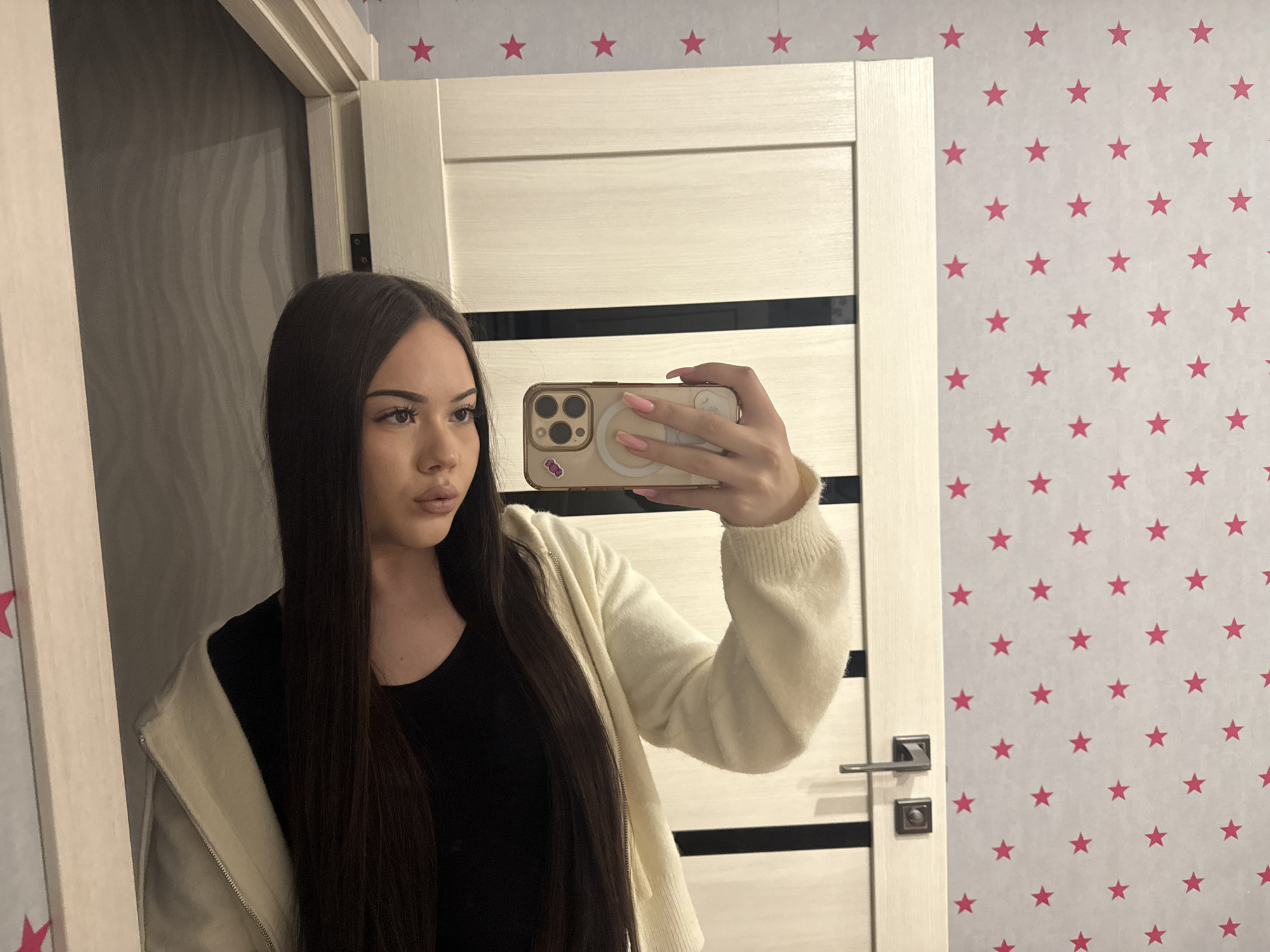 A young woman with long, straight dark hair takes a mirror selfie in an indoor setting, likely a private residence in Naberezhnye Chelny, Russia. She is reflected from the waist up, holding a light-colored smartphone with her right hand, which displays manicured nails. She is wearing a black top under an off-white, zippered hooded cardigan or jacket. Her expression is neutral, and she gazes slightly to her right within the reflection.
The background visible in the mirror consists of a light-colored door with dark horizontal accents, a modern handle, and a lock. To the right of the door is a wall covered in light grey or off-white wallpaper adorned with a repeating pattern of small pink stars. To the left, a darker, possibly textured wall or furniture is visible. The lighting appears to be artificial, typical of an indoor environment. No specific time of day or weather is discernible. There is no legible text visible in the image.