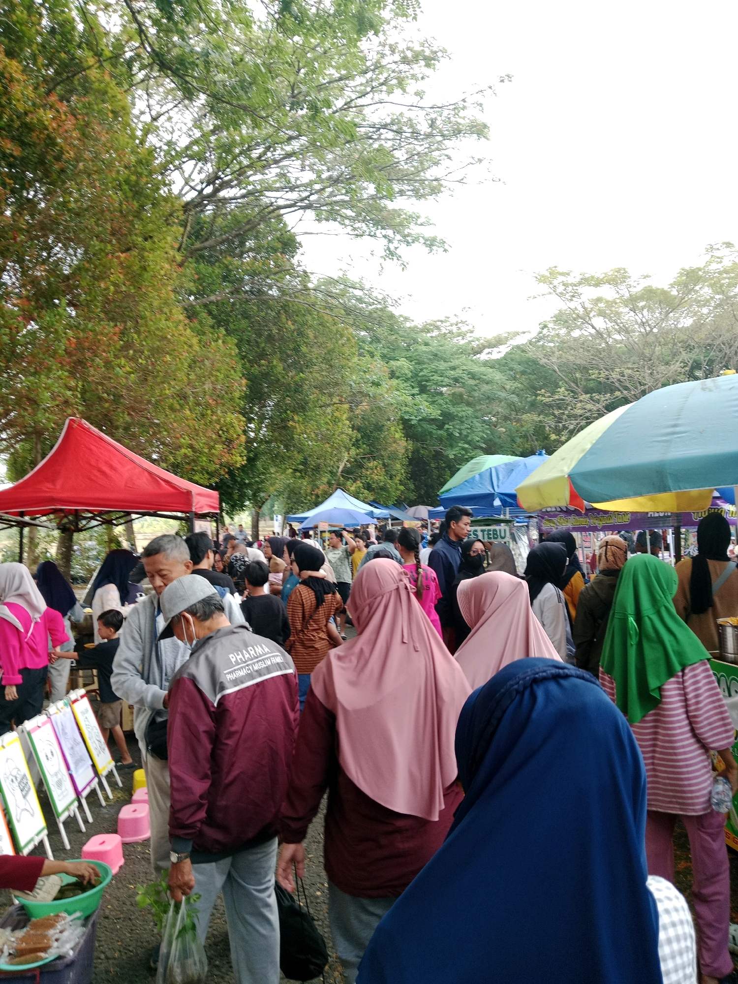This image depicts an outdoor market scene, likely in Ciamis, Indonesia, as suggested by the context. The foreground is dominated by a diverse group of people wearing various types of headscarves and modest clothing, indicating a Muslim-majority population. Several individuals are seen from the back, looking towards stalls and other shoppers. In the midground, there are numerous market stalls covered by colorful umbrellas and canopies, including a prominent red tent and a blue and green umbrella. These stalls display various goods, though the specifics are not entirely clear. The background features trees with lush green foliage, suggesting a park-like or natural setting within the city. The sky is overcast and bright. Some people appear to be browsing or interacting at the stalls. The ground appears to be gravel or dirt. The overall atmosphere suggests a bustling community event or market day.