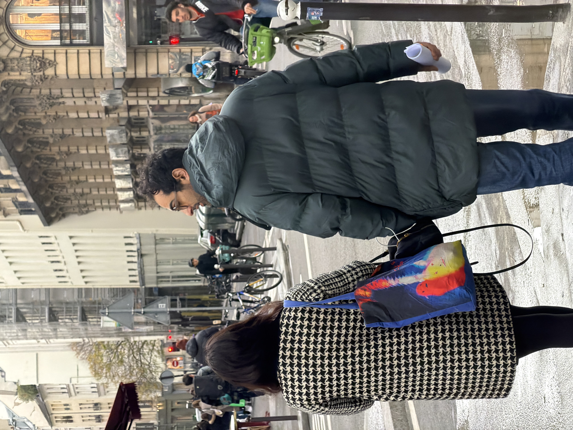 The image captures a street scene in Paris, France. The perspective is from behind two individuals, a man and a woman, walking on a sidewalk. The man is wearing a dark green puffer jacket, blue jeans, and glasses and carries a rolled-up white object in his right hand. The woman is wearing a houndstooth patterned jacket, black leggings, and carrying a bag with a colorful design. In the background, there are buildings with ornate facades, typical of Parisian architecture. Several bicycles and scooters are parked along the street, and people can be seen riding them. Traffic lights are visible in the distance. The ground appears wet, suggesting it may have recently rained. Overall, the image portrays a typical day in a Parisian urban setting.