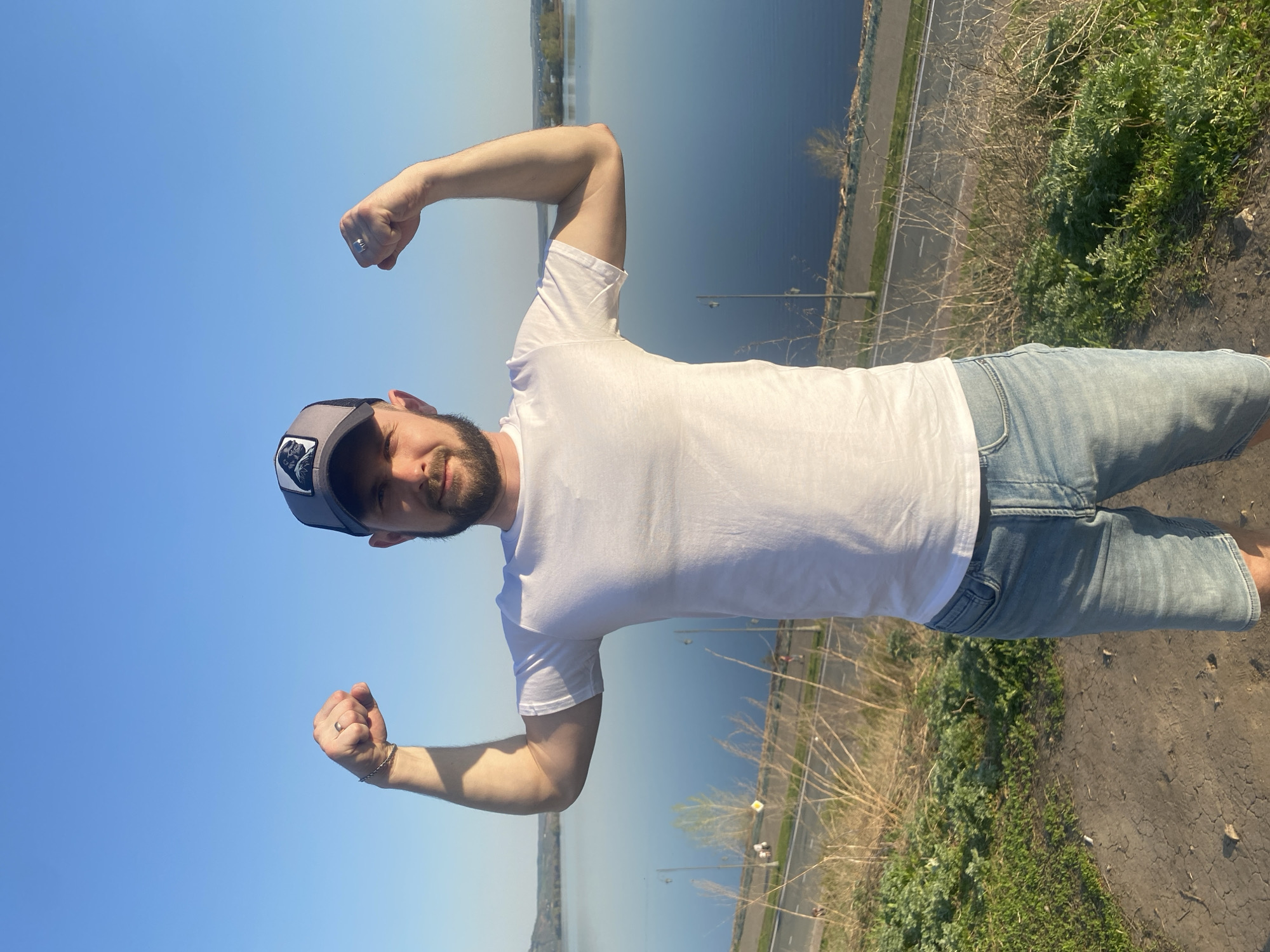 A man stands near water, flexing his arms. He wears a white t-shirt, denim shorts, and a baseball cap. The location appears to be outdoors in Sviyazhsk, Russia, adjacent to a body of water with a visible shoreline and road with streetlights and possible cyclist present. There is vegetation on the ground near where he stands. The sky is mostly clear.