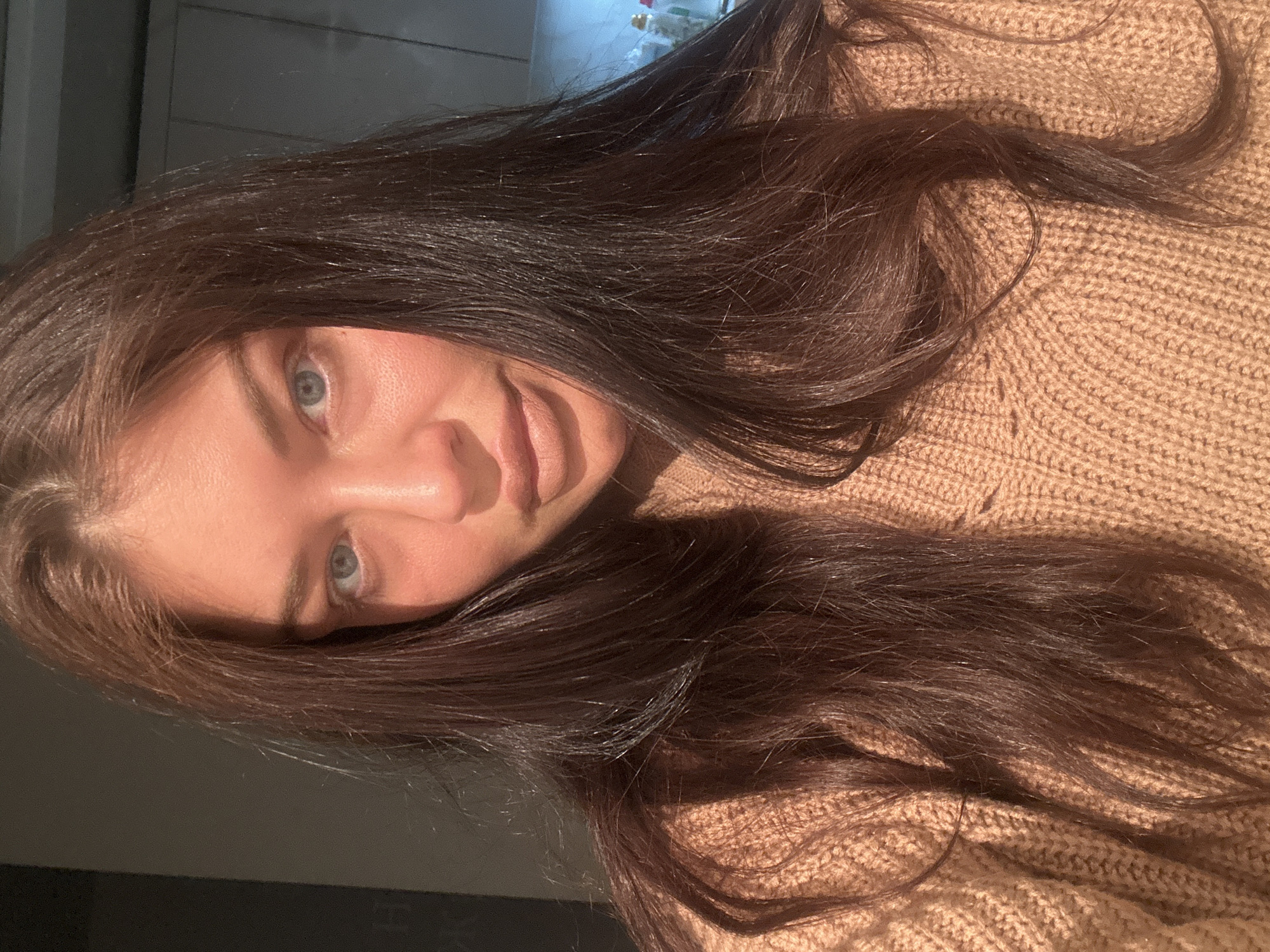 A woman with long, dark hair and blue eyes is facing the camera. She is wearing a beige knit sweater. The lighting is somewhat dim, casting shadows across her face. The background is out of focus, revealing a blurred area with some objects that might be bottles or jars. It is impossible to determine if this image was taken in Khimki, Russia or anywhere else based on the provided visual information.