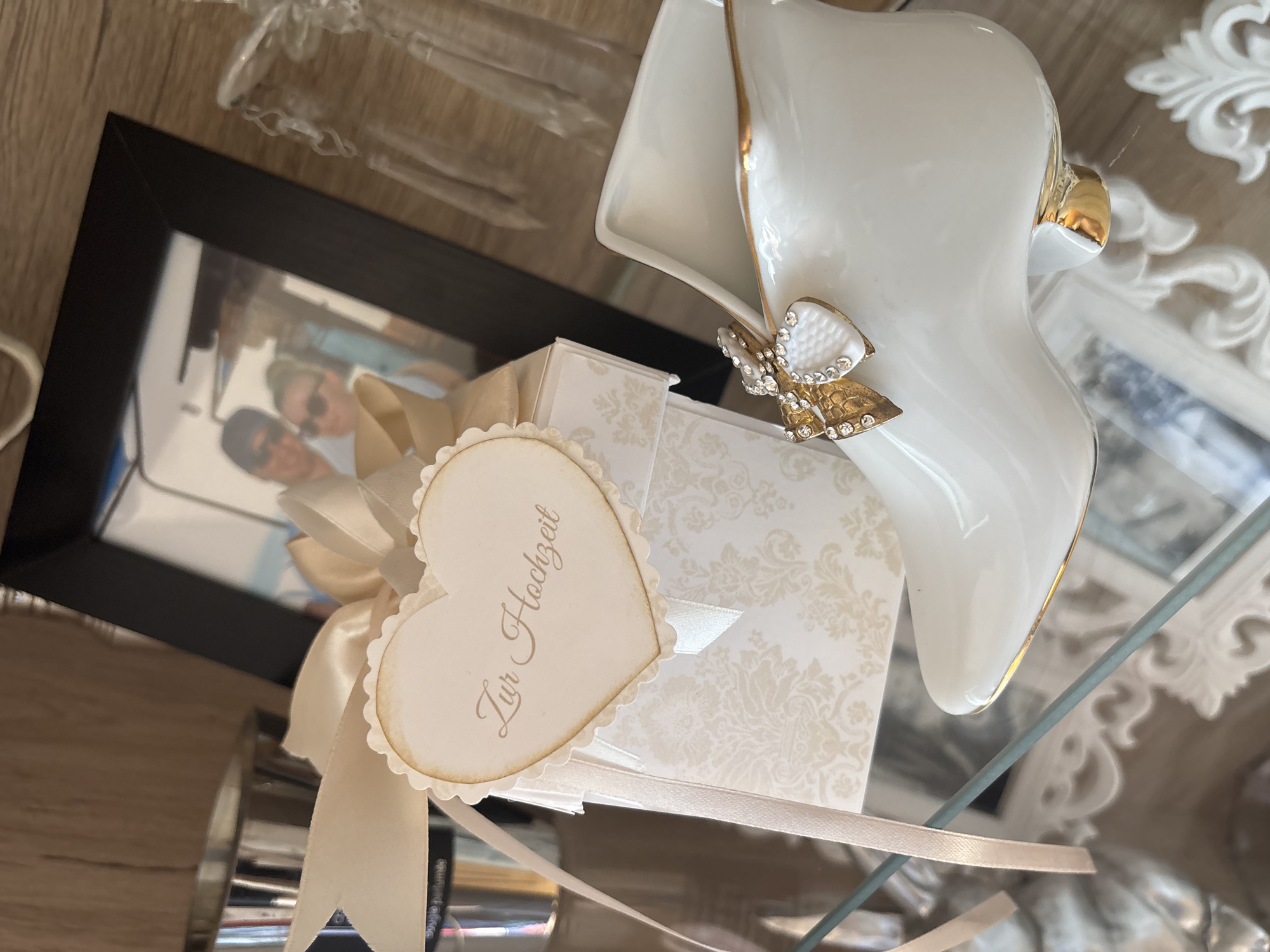 An indoor scene showcases a collection of elegant, wedding-themed decorative items arranged on a reflective glass surface. The central focus is a square gift box, rendered in light cream with an intricate damask pattern, secured with a matching satin ribbon. A heart-shaped tag, also cream with scalloped edges, is tied to the ribbon, bearing the German inscription "Zur Hochzeit" (To the Wedding).
Adjacent to the gift box, on the right, stands a pristine white ceramic decorative object shaped like a high-heeled shoe or boot. It features delicate gold trim along its contours and is adorned with a prominent golden bow embellished with sparkling rhinestones on its side.
Behind the gift box, a black-framed photograph is visible, tilted to the left. The image within the frame shows a couple, a man and a woman, both wearing sunglasses, suggesting a bright, outdoor setting and a relaxed atmosphere. Other elements in the background include a partially visible clear, possibly crystal, decorative item in the upper left, and reflections on the glass surface hinting at ornate white decor and a wooden texture. The overall aesthetic, dominated by whites, creams, and gold, conveys a celebratory and elegant mood, indicative of a wedding event or cherished marital memories.