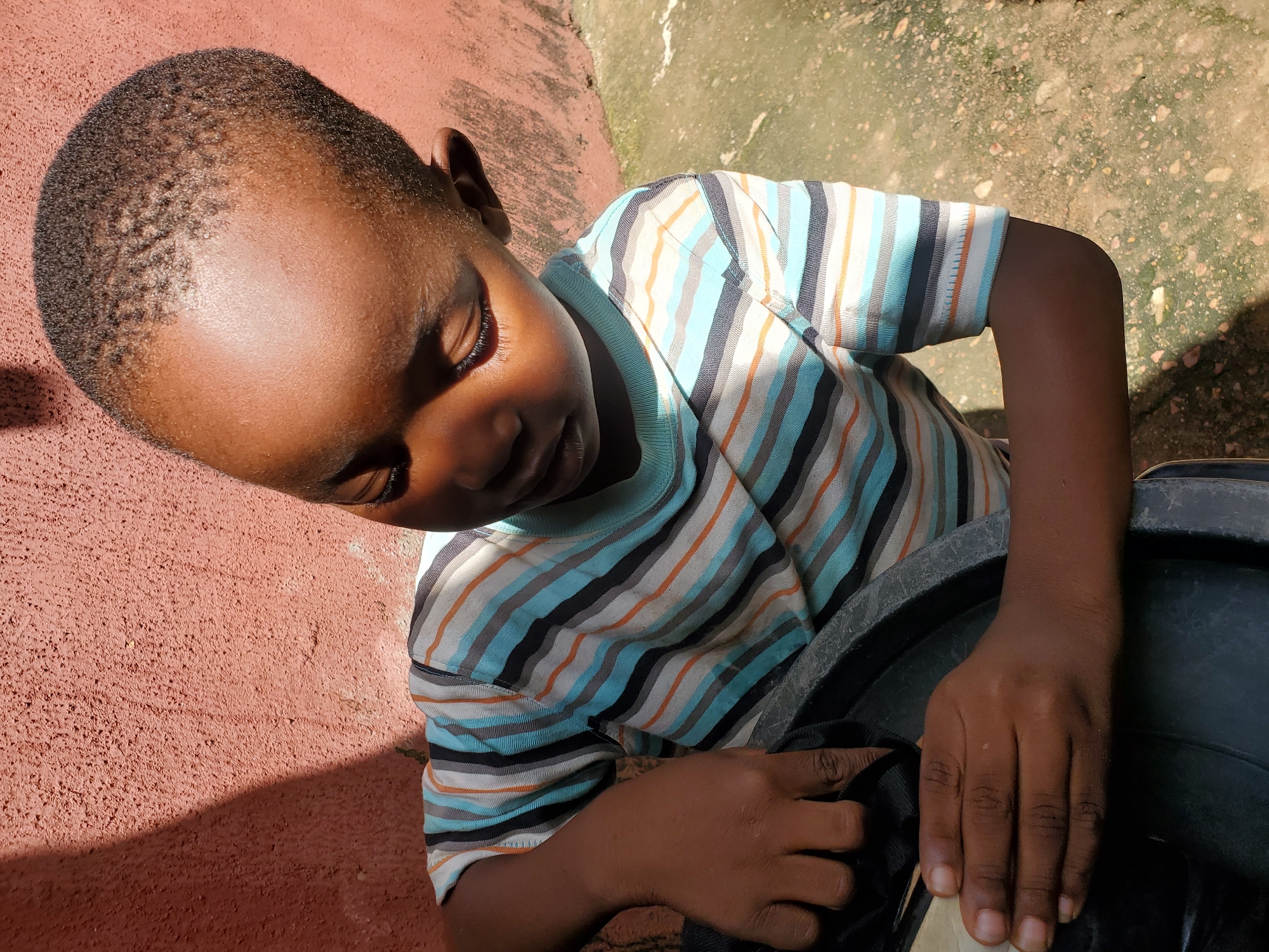 The image shows a close-up view of a young Black child from the shoulders up, looking downwards with their eyes closed. The child is wearing a horizontally striped t-shirt with colors including blue, white, orange, and black. The child's left arm is extended and their hands are resting on the rim of a dark-colored, possibly plastic, container. The background consists of a textured, reddish-pink wall on the left and a mottled, greenish-brown ground surface on the right, with scattered pebbles. Sunlight casts a shadow across the left side of the image, highlighting the child's head and right shoulder. The scene appears to be outdoors, possibly in a residential or public area, with the lighting suggesting it is daytime. The location is identified as the city of Jos, Nigeria.